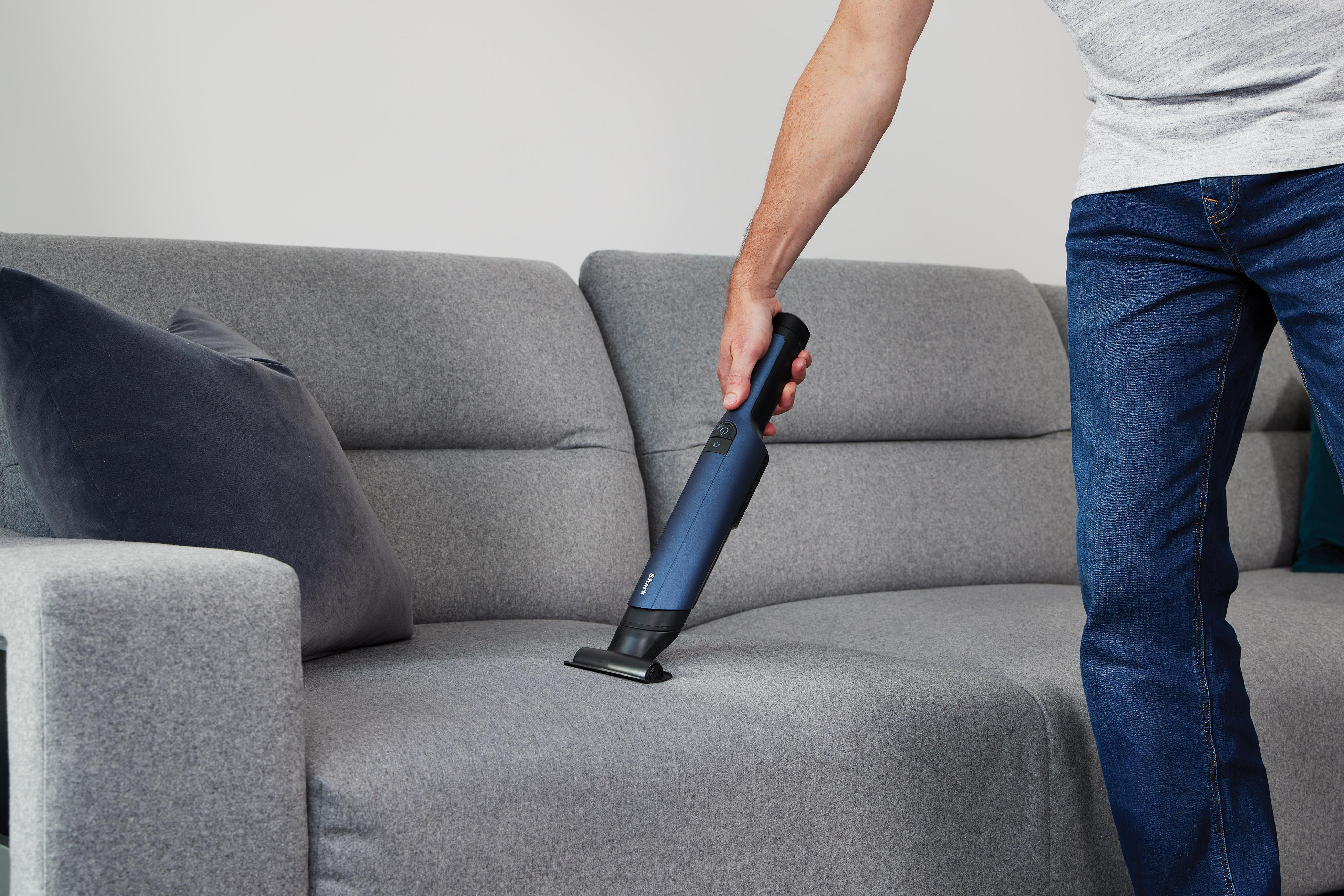 Ein Mann saugt ein graues Sofa mit einem Handstaubsauger. Er trägt Jeans und ein graues Hemd.