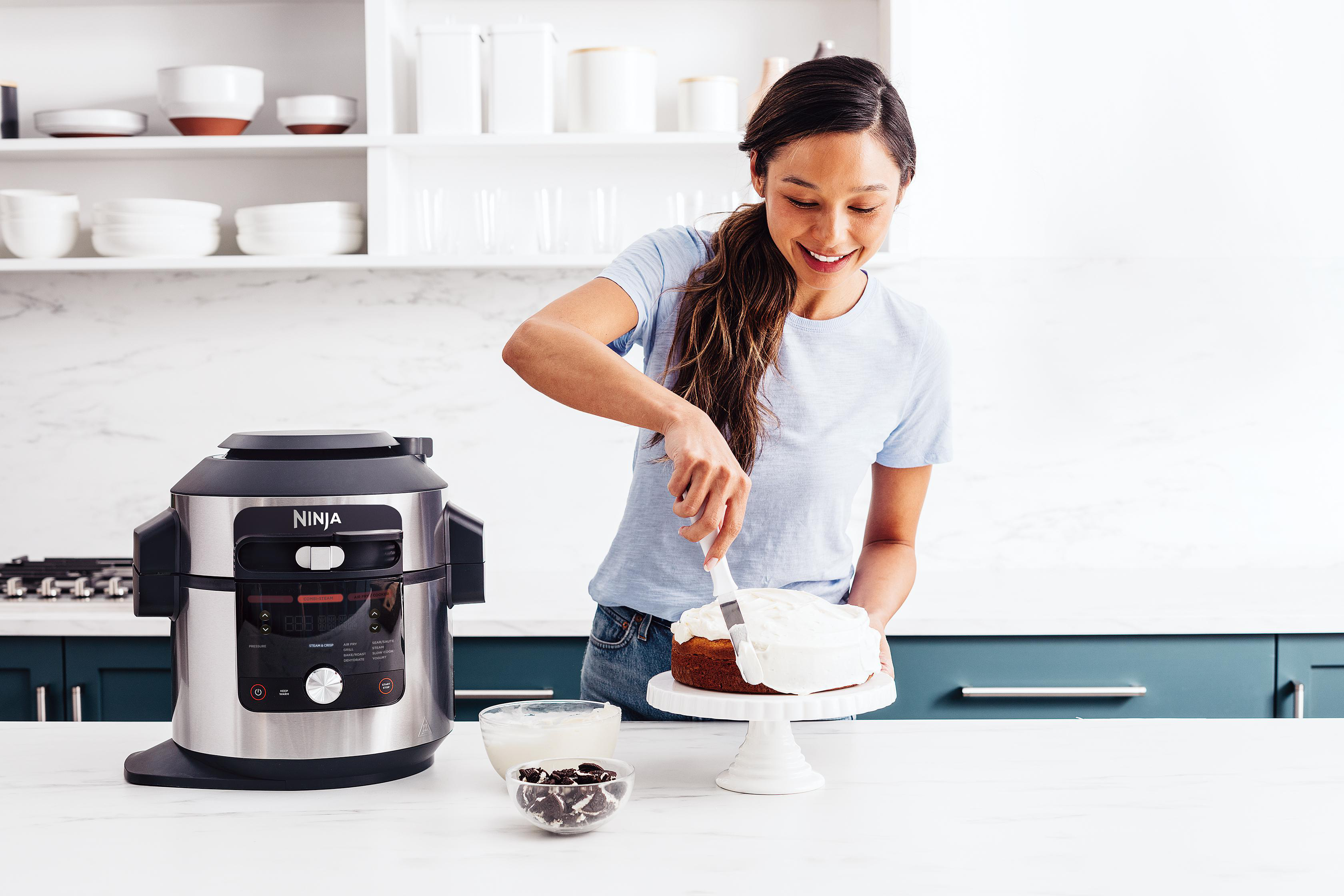 Eine Frau, die einen Kuchen auf einem Ständer mit dem Ninja Foodi-Gerät und Zutaten in der Nähe verziert.