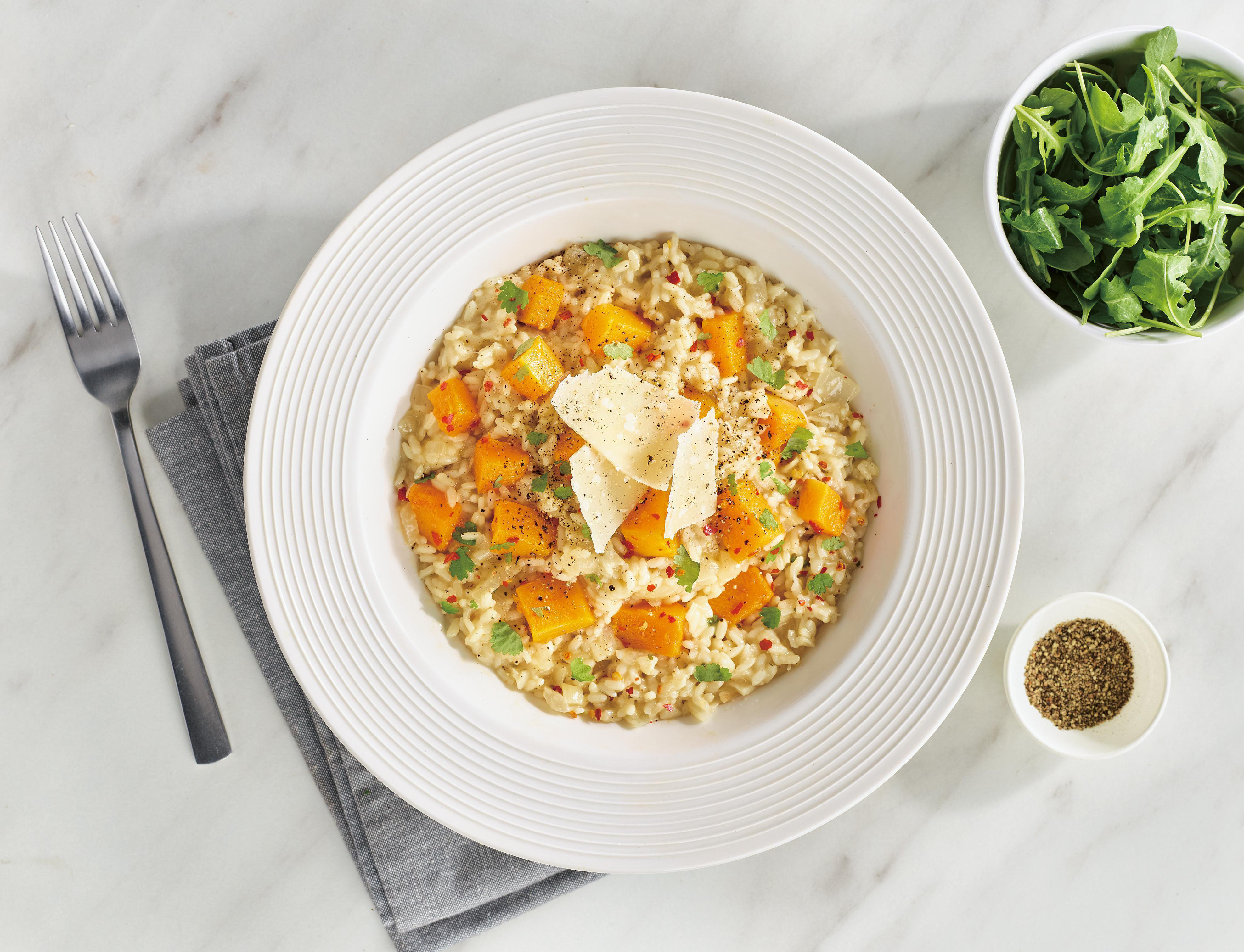 Eine weiße Schüssel mit Risotto und Butternusskürbis, garniert mit Käse und Pfeffer.