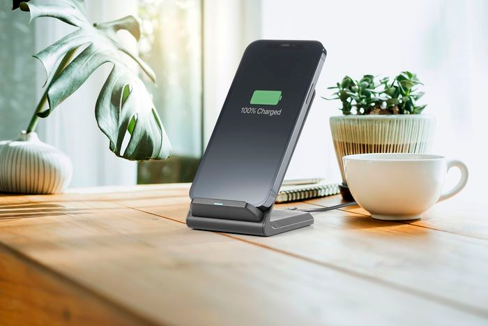 Smartphone sur un chargeur sans fil, affichant 100% chargé. Une plante et une tasse à café sont à proximité sur une table.