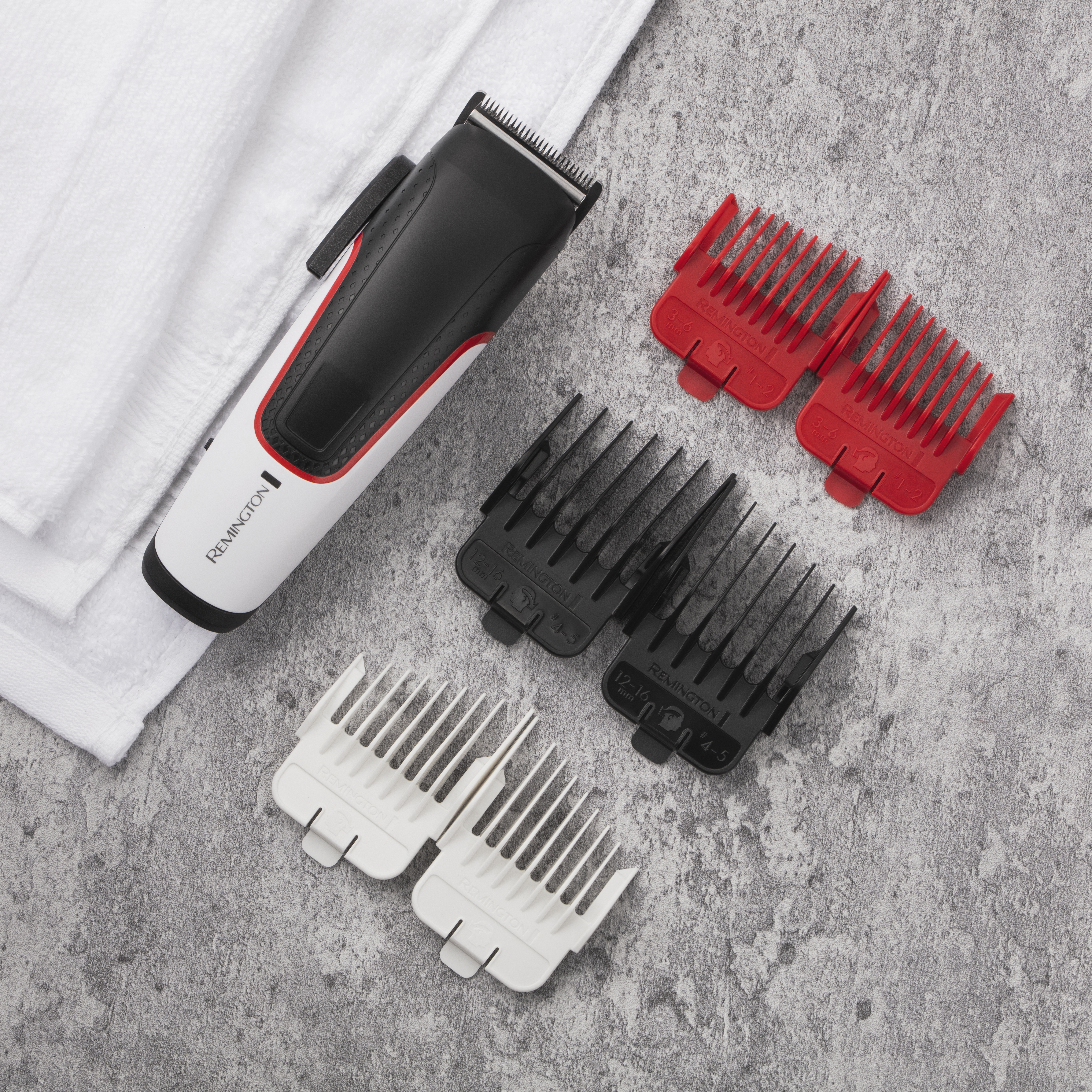 Haarclippers, rode, zwarte en witte kammen en een witte handdoek op een grijs oppervlak.