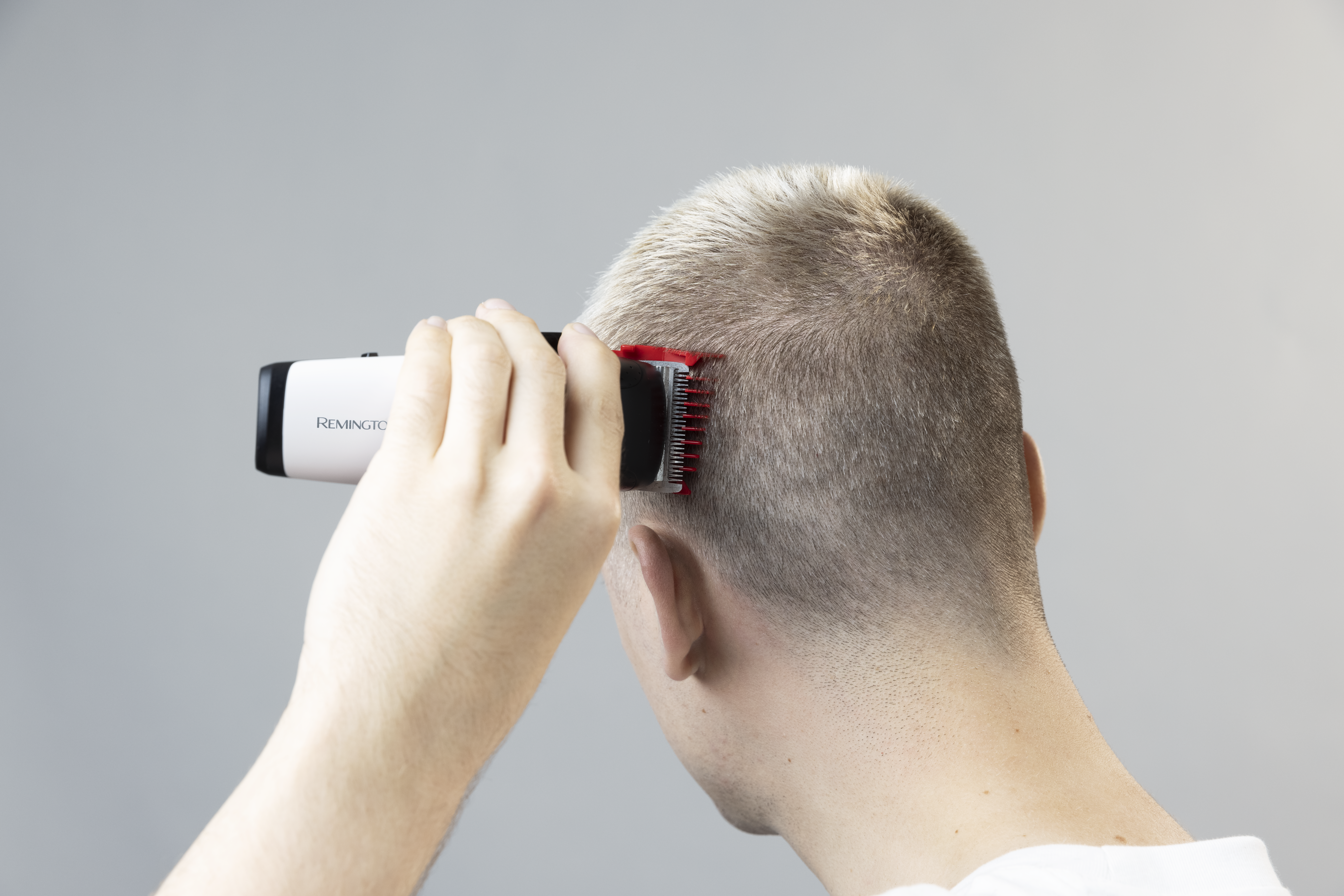 Een persoon knipt zijn haar met een Remington tondeuse. De tondeuse is rood en zwart. De achtergrond is een effen grijze muur.