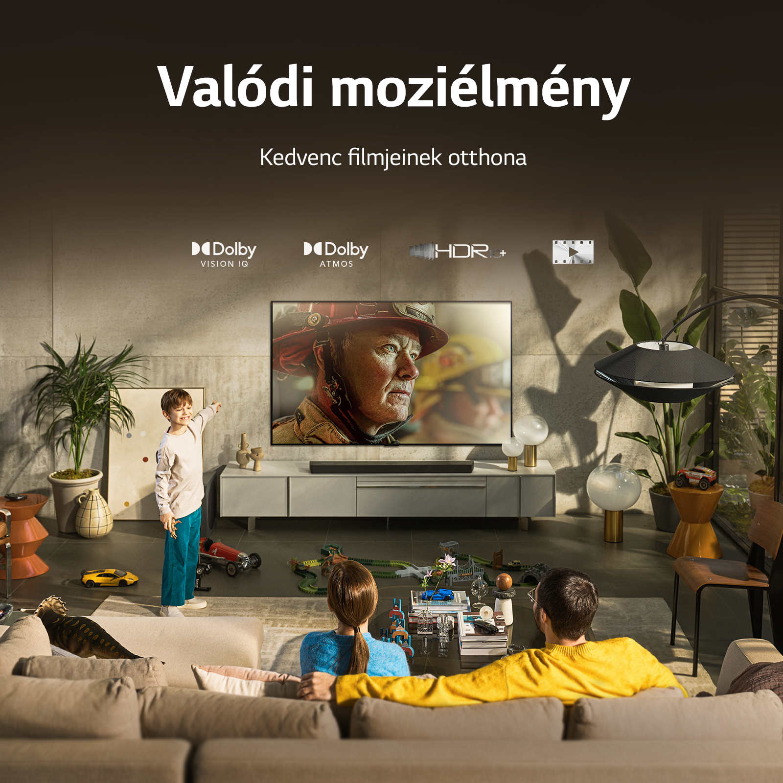 Nappali TV-vel. Egy fiú a képernyőre mutat; egy pár a kanapén ül.