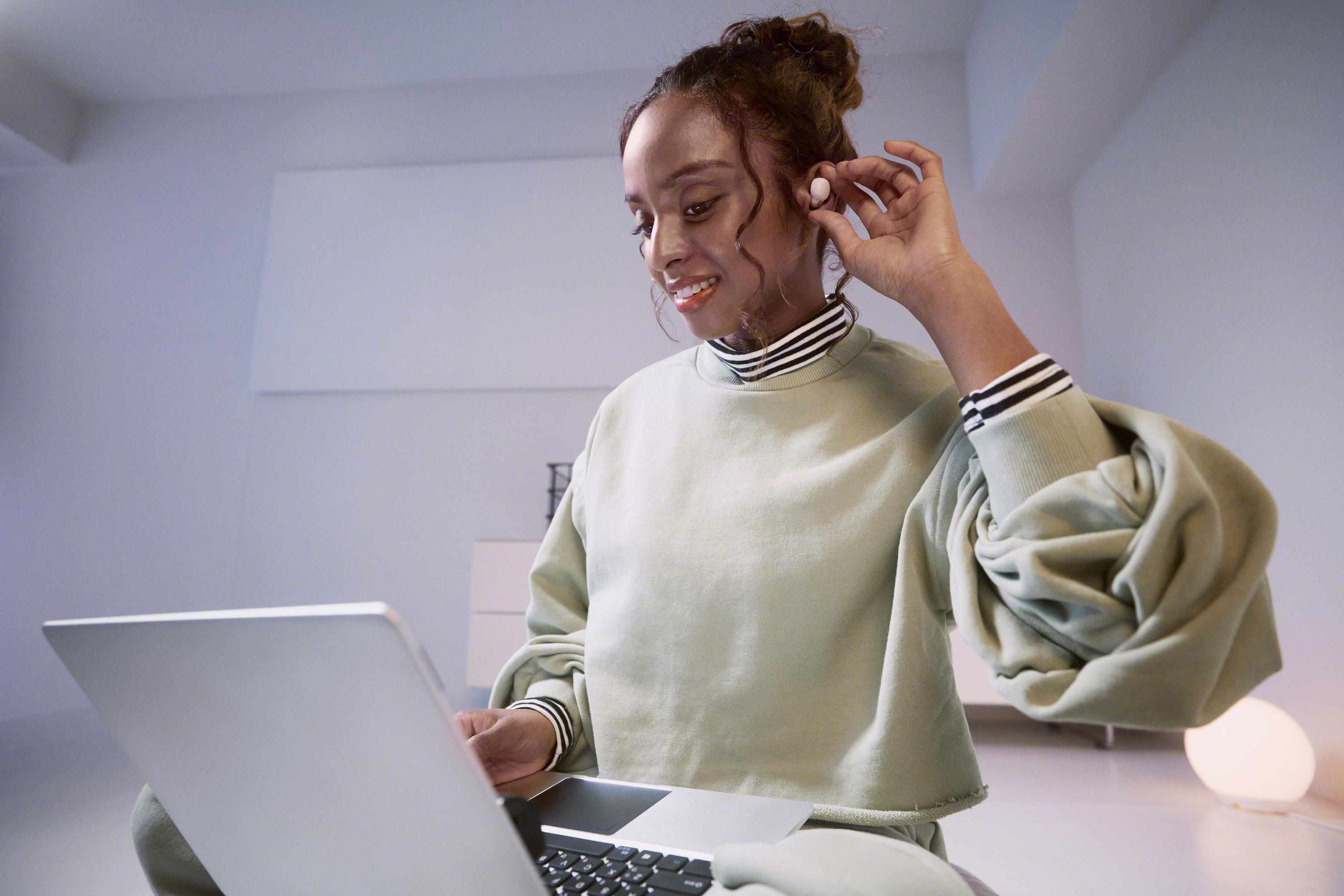 Eine Frau mit grünem Pullover benutzt einen Laptop, während sie einen Ohrhörer anpasst.
