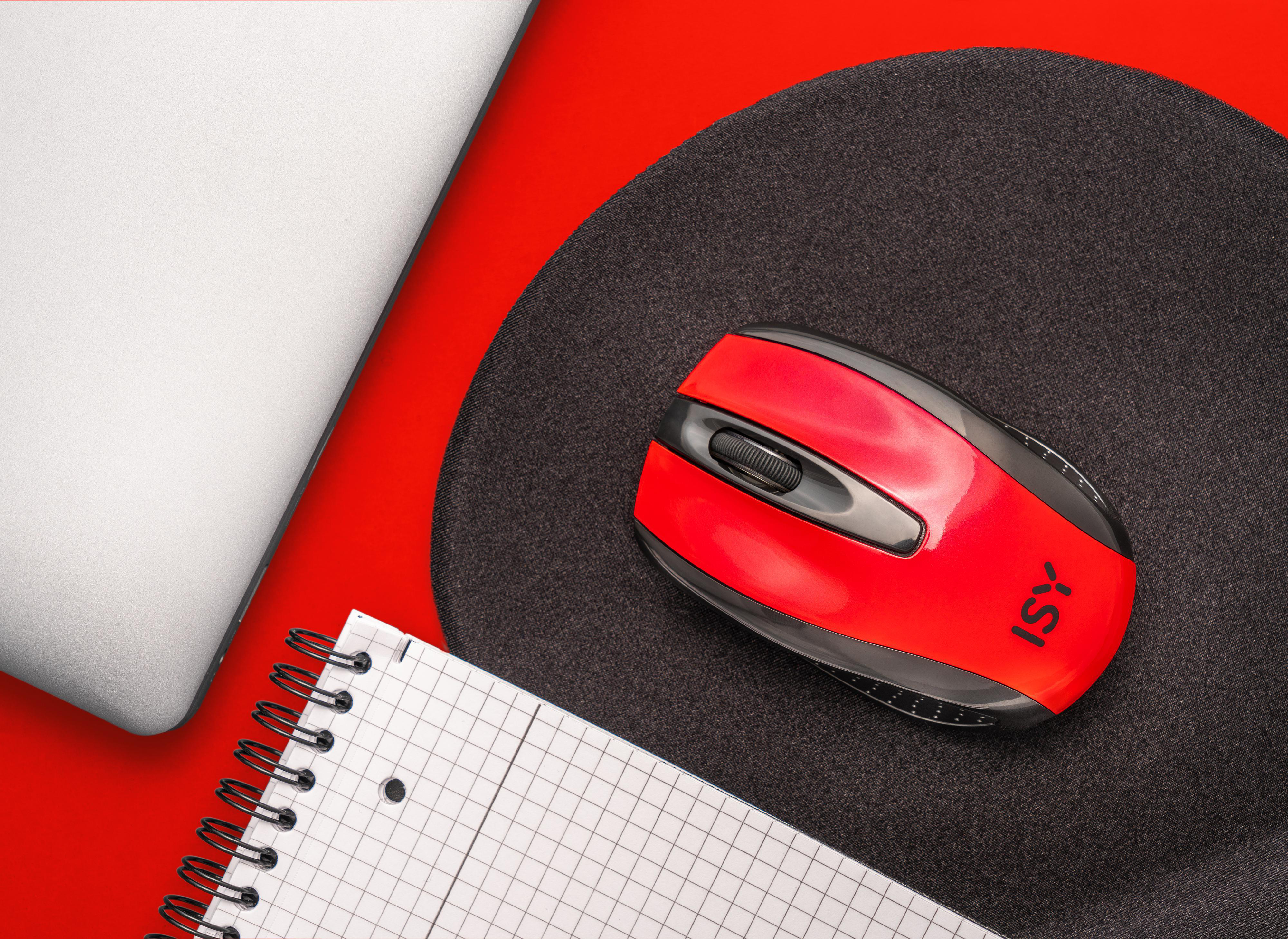 Eine rote Maus auf einem schwarzen Mauspads, mit Laptop und Notizblock auf roter Oberfläche.