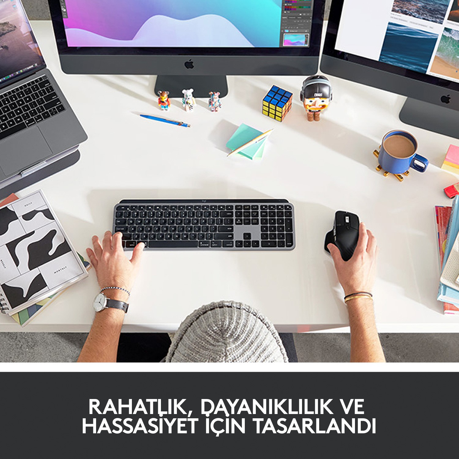 Eller beyaz bir masada klavye ve fare kullanıyor, monitörler ve aksesuarlar var.