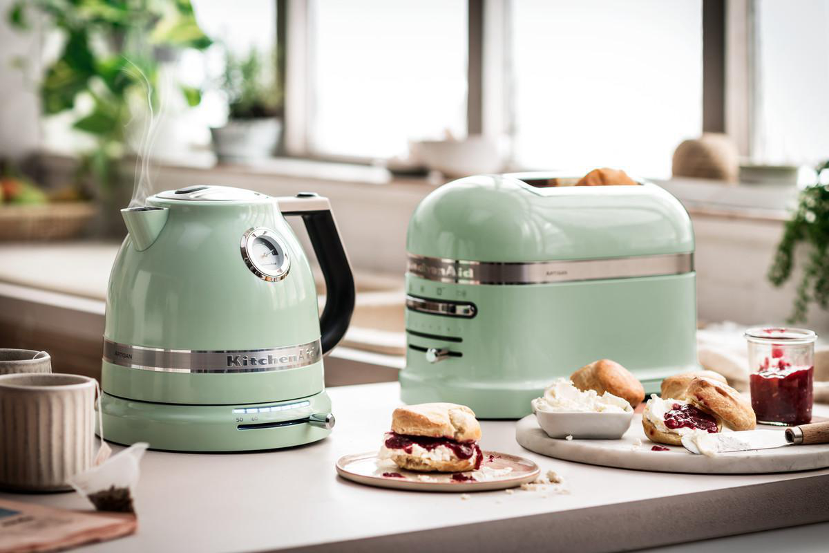 Ein mintgrüner KitchenAid Wasserkocher und Toaster mit Scones, Marmelade und Tee auf weißer Oberfläche.