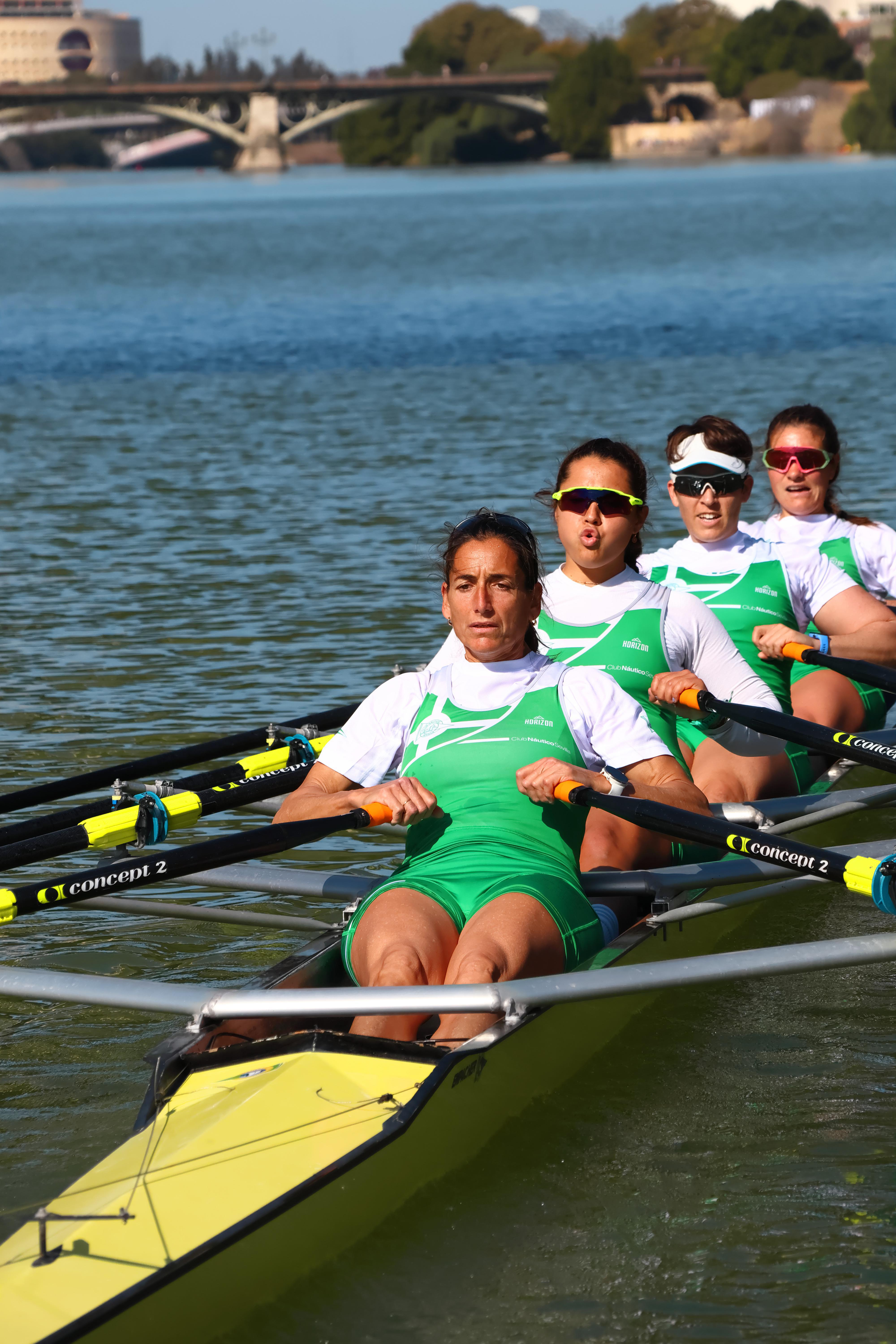 Vier Ruderer in einem Boot auf einem See. Grüne Uniformen, gelbes Boot, Gebäude im Hintergrund.