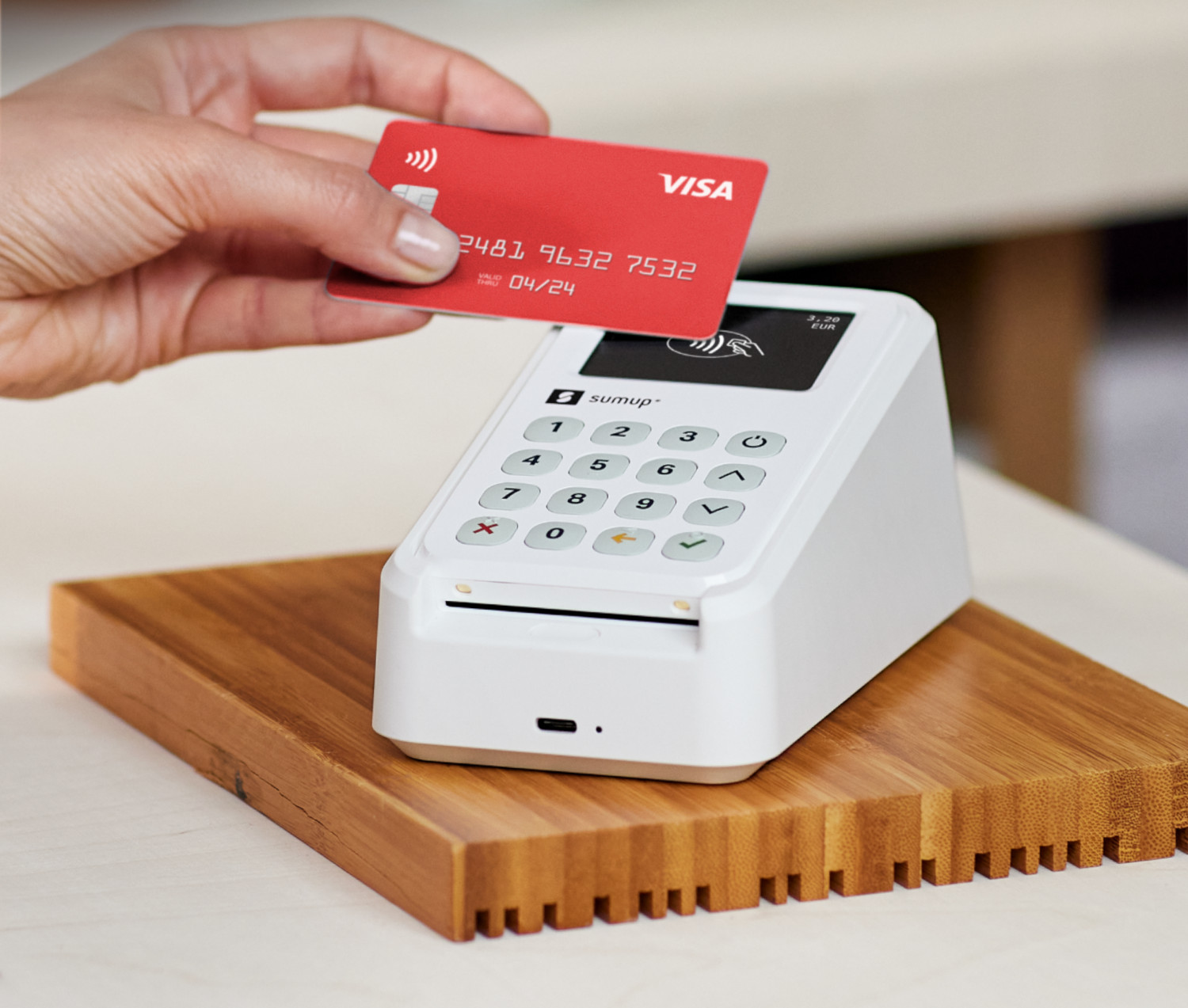 Une main place une carte Visa rouge au-dessus d'un lecteur de carte SumUp blanc sur une base en bois.