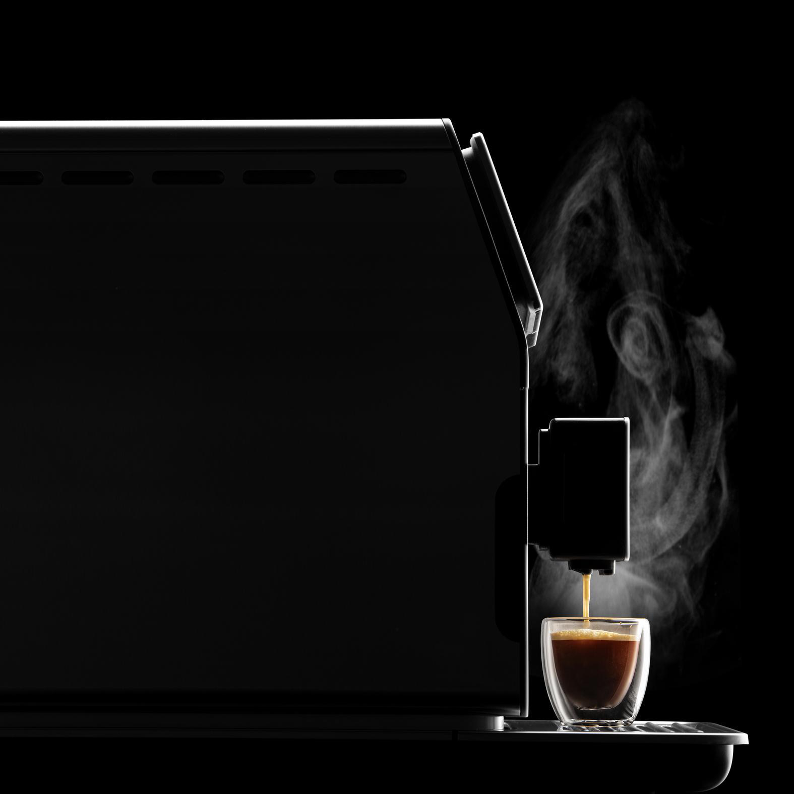 Schwarze Kaffeemaschine. Kaffee gießt in eine Glastasse. Dampf steigt vor schwarzem Hintergrund auf.