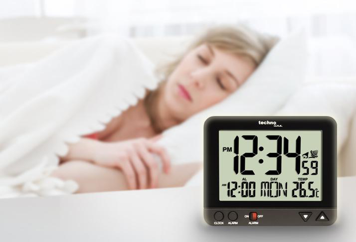 Eine Digitaluhr auf einem Tisch mit einer schlafenden Frau im Hintergrund.