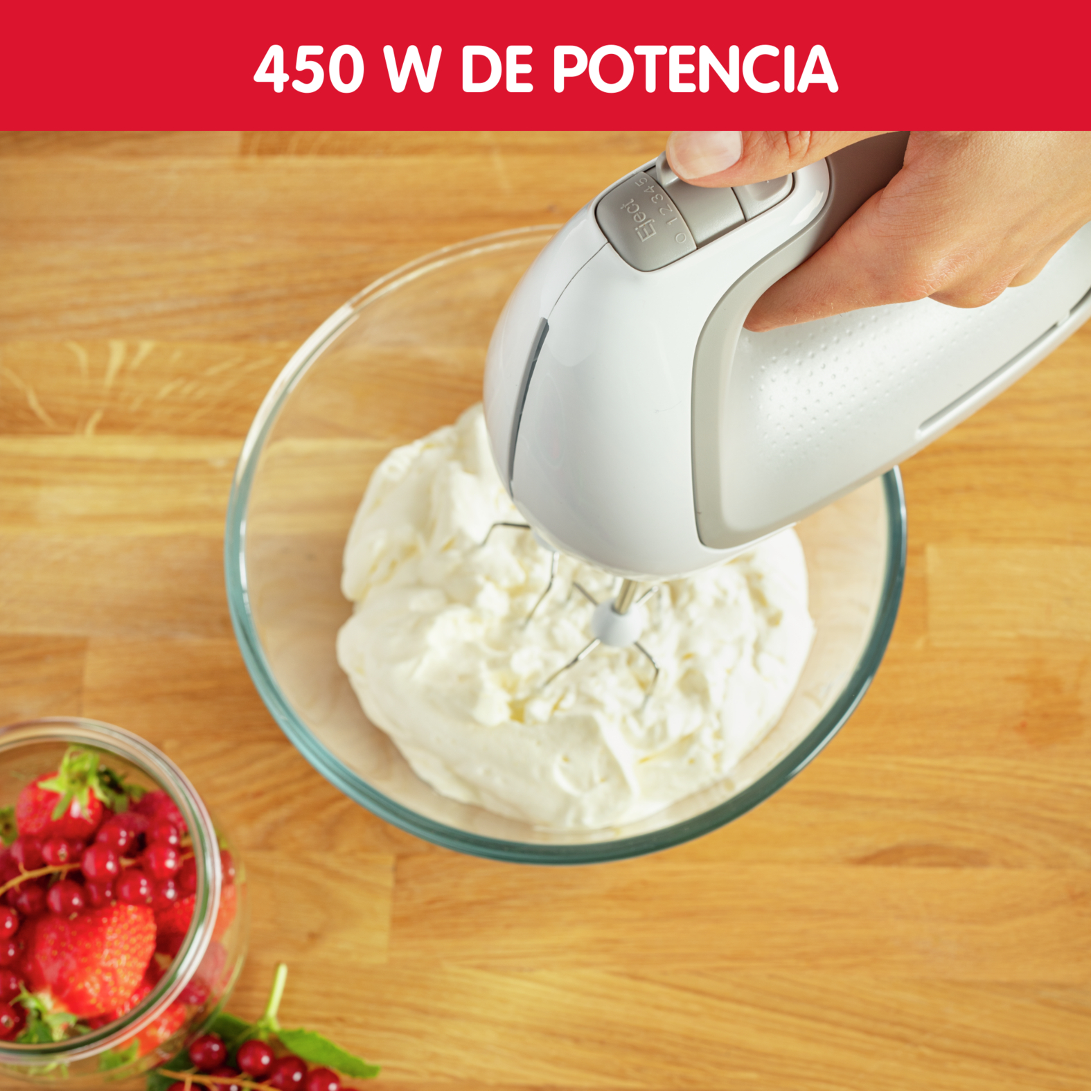 Batidora de mano mezclando crema en un bol, fresas y grosellas rojas a un lado.