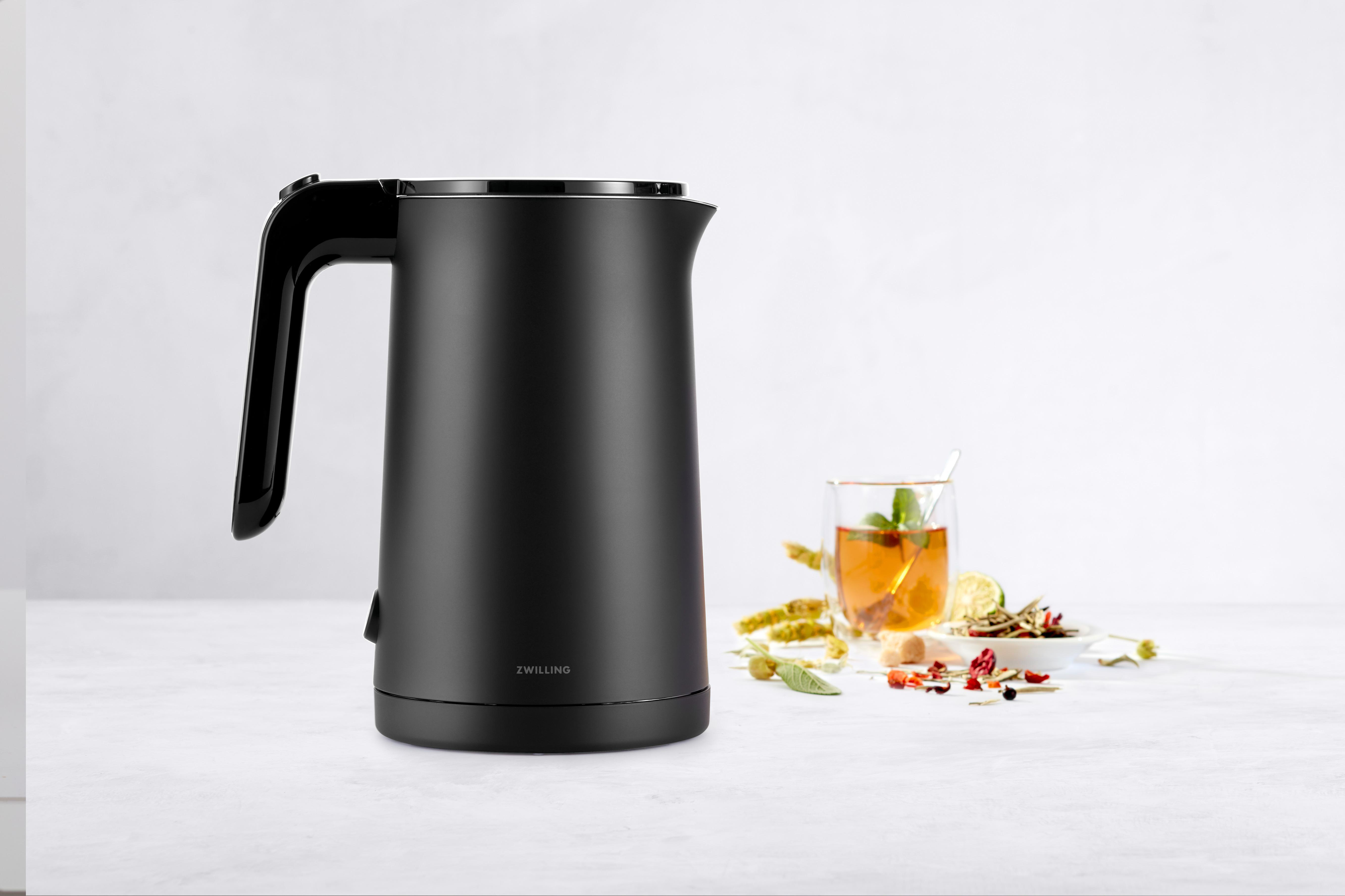 Schwarzer Wasserkocher mit Griff und ein Glas Tee auf einem Tisch.