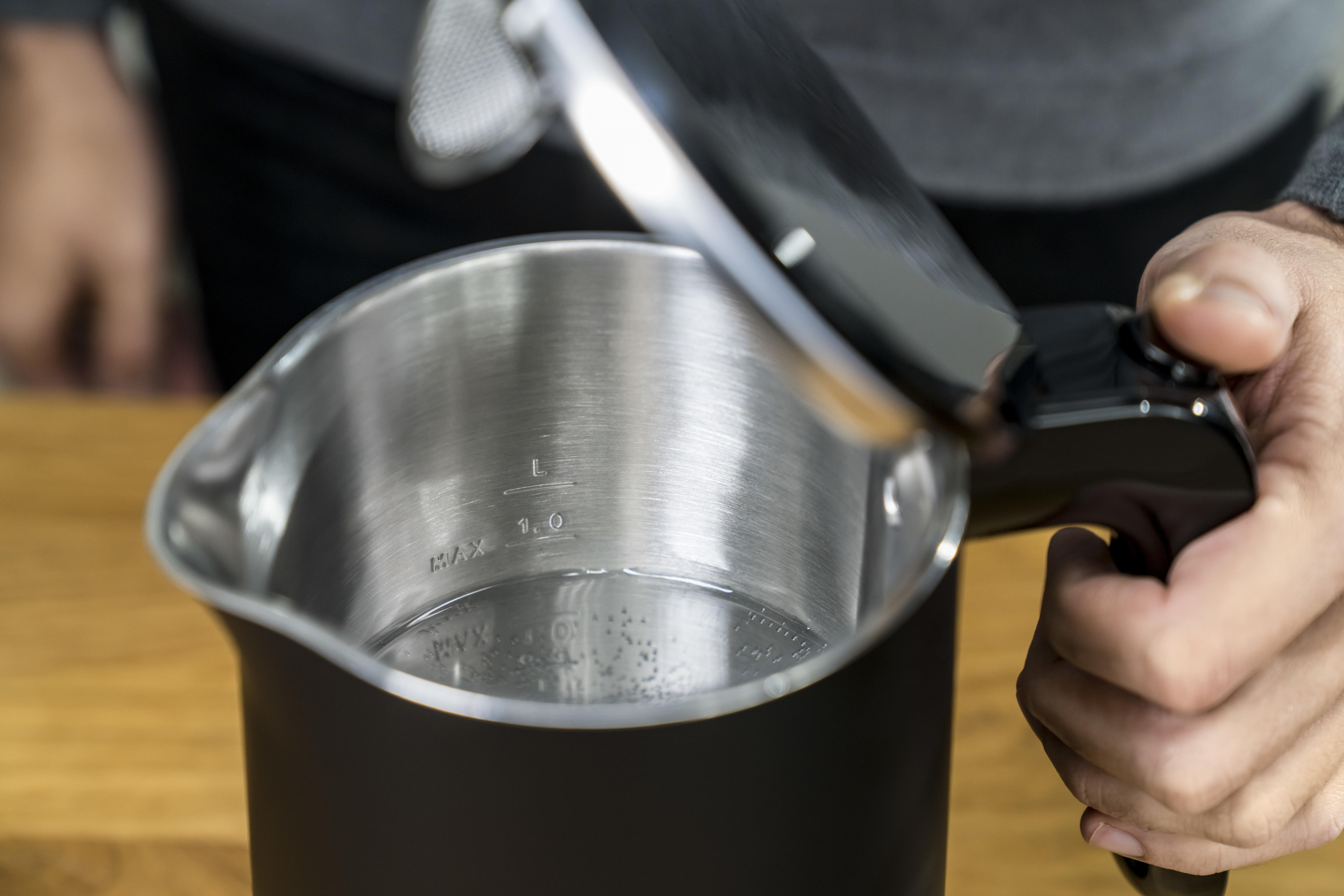 Schwarzer Wasserkocher mit Wasser, offener Deckel, Markierungen zeigen 1,0 l max. Holztisch sichtbar.
