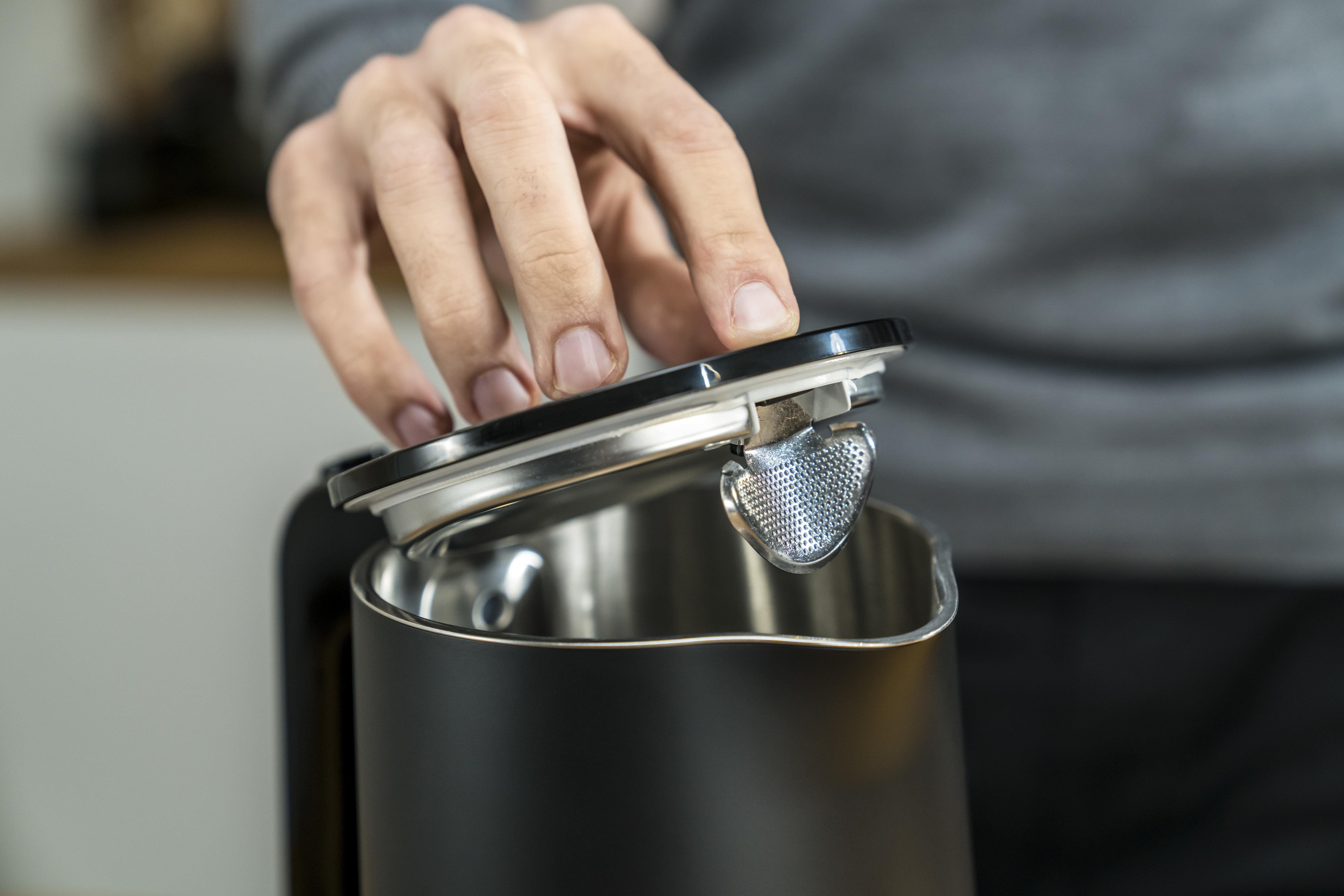 Eine Hand öffnet den Deckel eines schwarzen Wasserkochers und gibt einen Filter frei. Küchenumgebung.