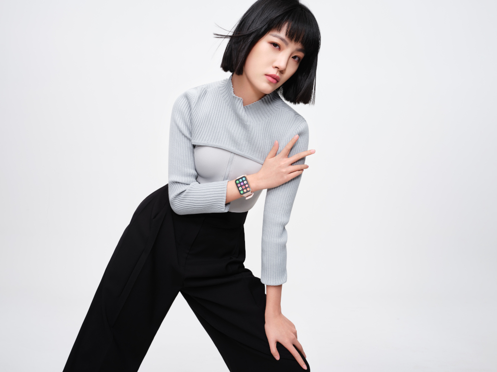Femme en pull gris clair et pantalon noir, porte une montre connectée, posant en studio.
