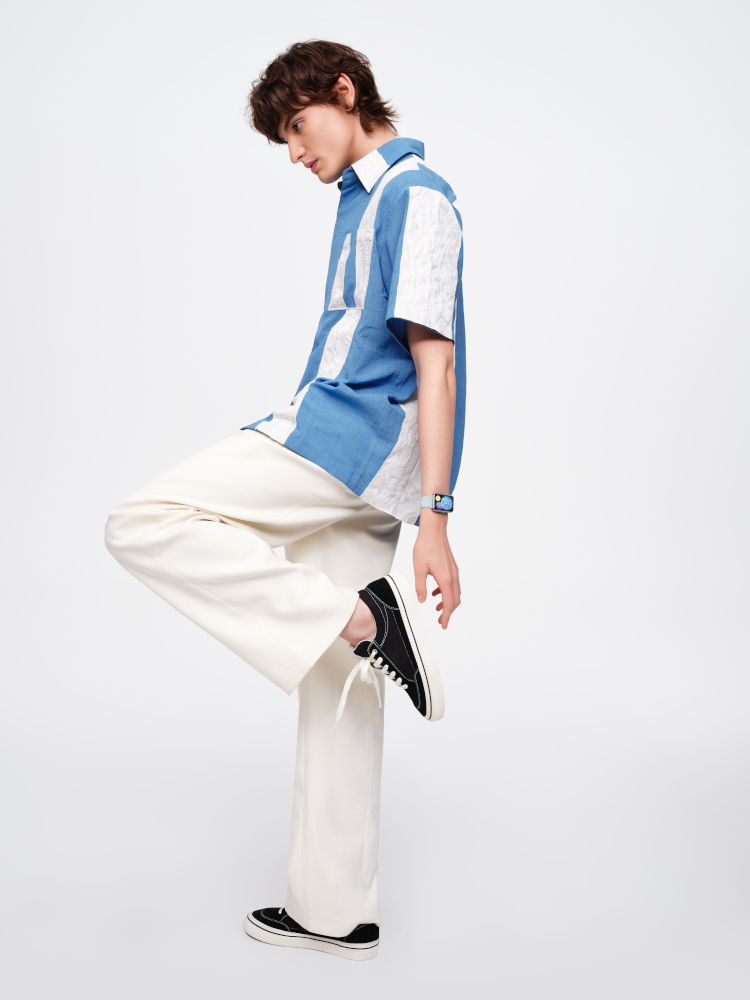 Un homme en pantalon blanc et chemise bleue et blanche, portant des baskets noires, debout sur une jambe.