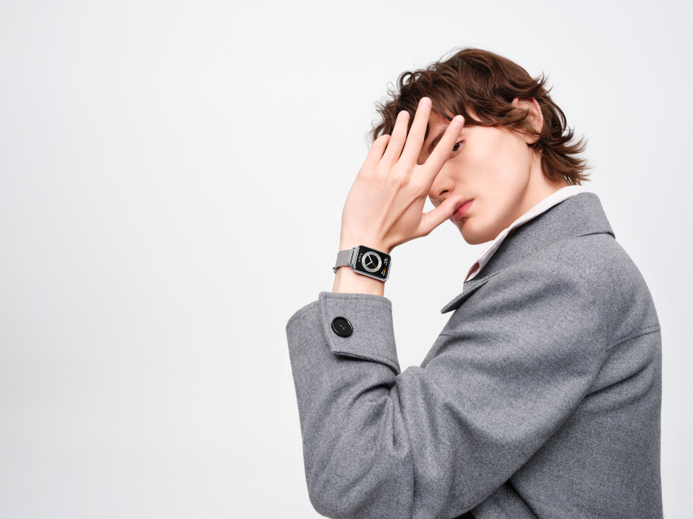 Un homme se protégeant le visage avec sa main, portant une montre et un manteau gris, fond blanc.