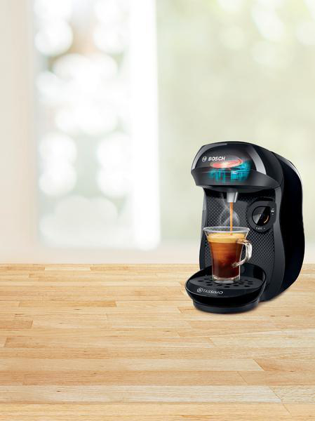 Eine schwarze Kaffeemaschine mit einem klaren Glas Kaffee. Ein Holztisch und ein Fenster im Hintergrund.