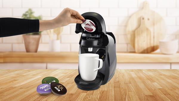 Hand legt Kaffeepad in schwarze Kaffeemaschine. Weiße Tasse darunter. Holzoberfläche.