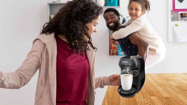 Frau hält eine Tasse Kaffee, während sie auf die Kaffeemaschine schaut, mit Familie im Hintergrund.