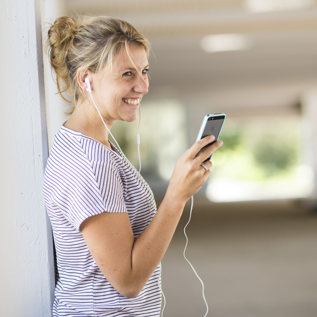 Blonde Frau, trägt gestreiftes Hemd, lächelt und hört Musik auf ihrem Handy mit Ohrhörern.