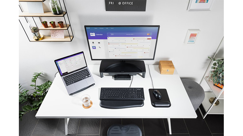 Egy otthoni irodai beállítás monitorral, laptop, billentyűzet, egér és asztali kiegészítőkkel egy fehér asztalon.