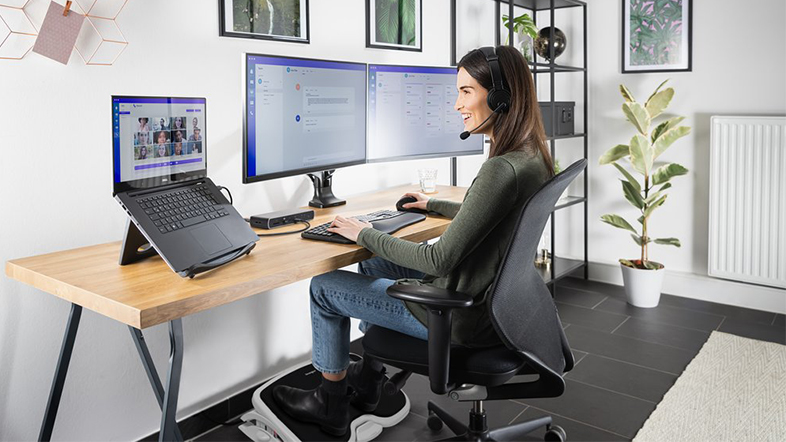 Egy nő laptopon dolgozik, ami két monitorhoz van csatlakoztatva, fejhallgatót visel.