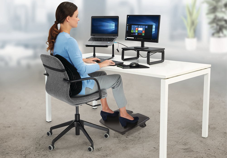 Nő számítógépet használ laptop, monitor, ergonomikus szék és lábtartó az asztalnál.