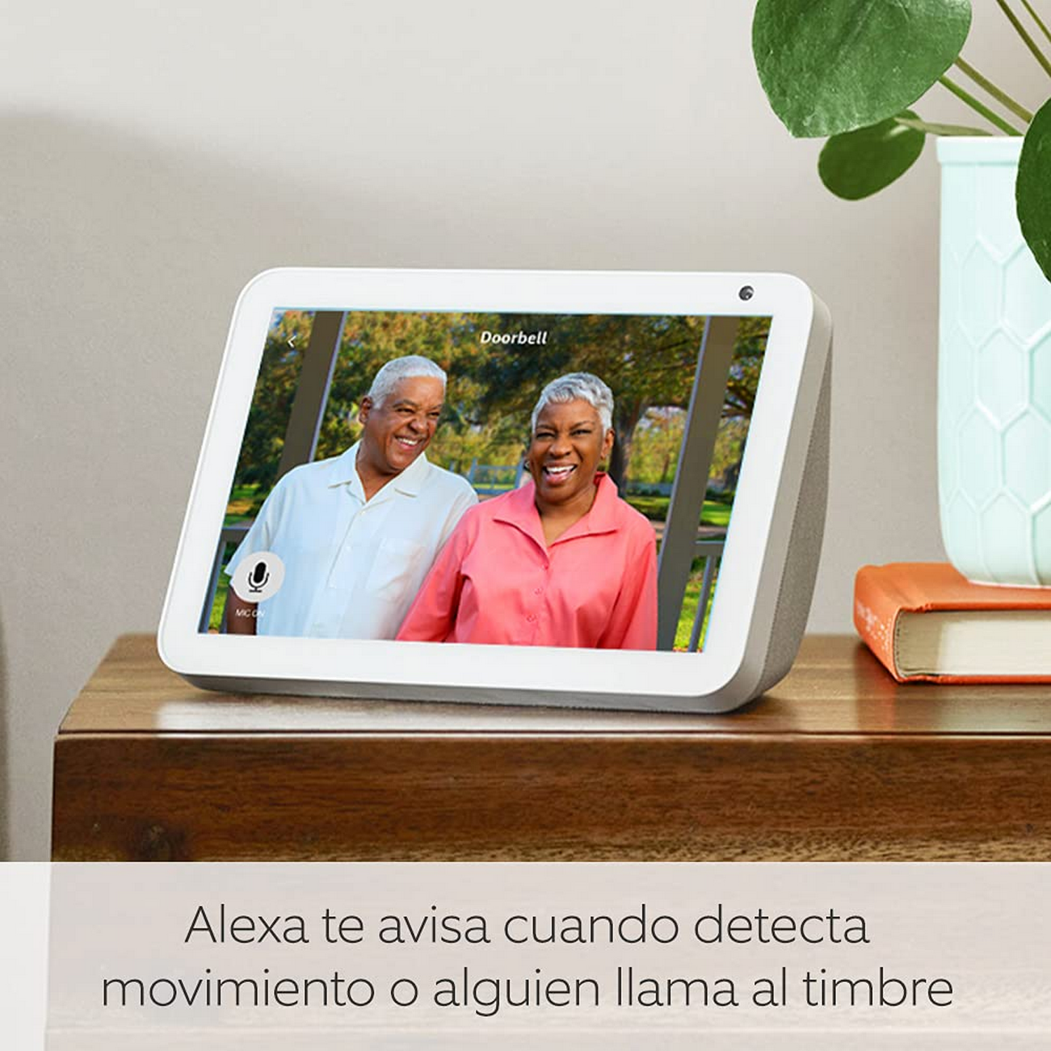Una pantalla inteligente muestra a una pareja de ancianos. El dispositivo está en una mesa de madera. Una planta al fondo.