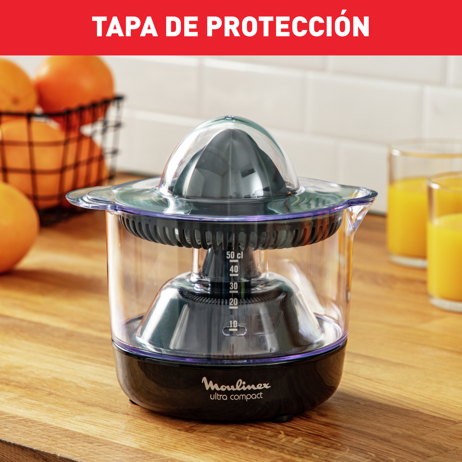 Una exprimidora con tapa etiquetada 'TAPA DE PROTECCIÓN'. Naranjas en una cesta en el fondo.