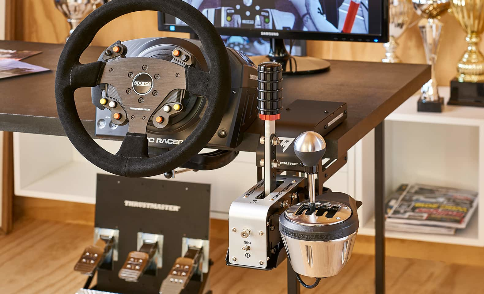 Un volante da corsa nero con cambio, pedali e monitor su una scrivania in legno.