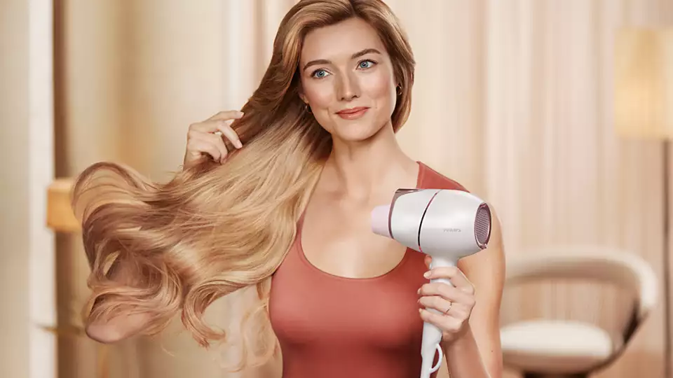 A woman with long blonde hair uses a white and pink hair dryer.
