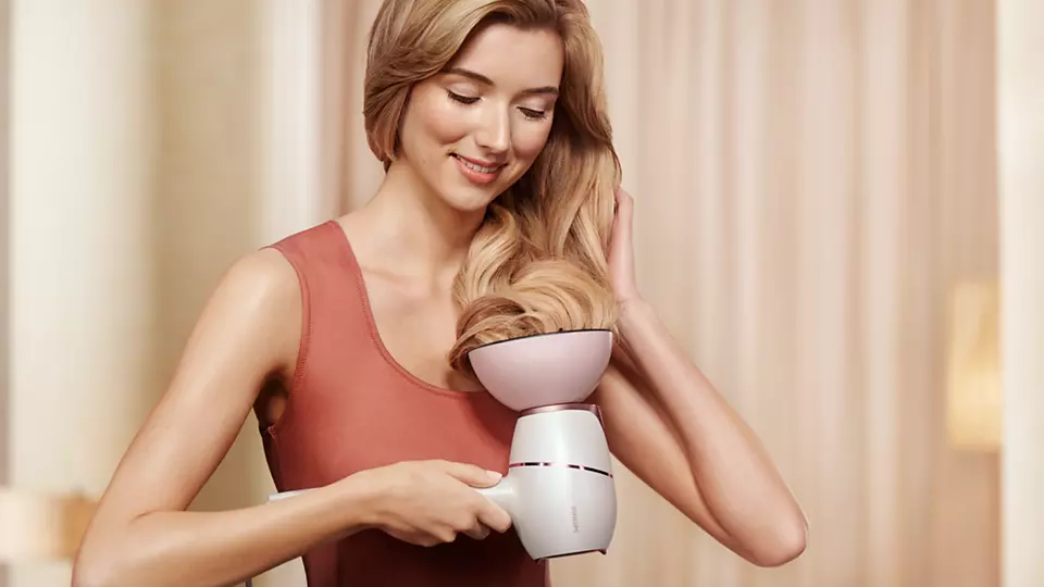 Woman drying hair with a hair dryer. She is in a room with a beige wall and a mirror.
