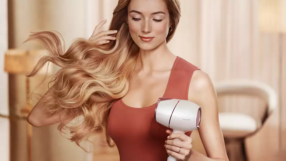 Woman styling her long blonde hair with a white and rose gold hair dryer.