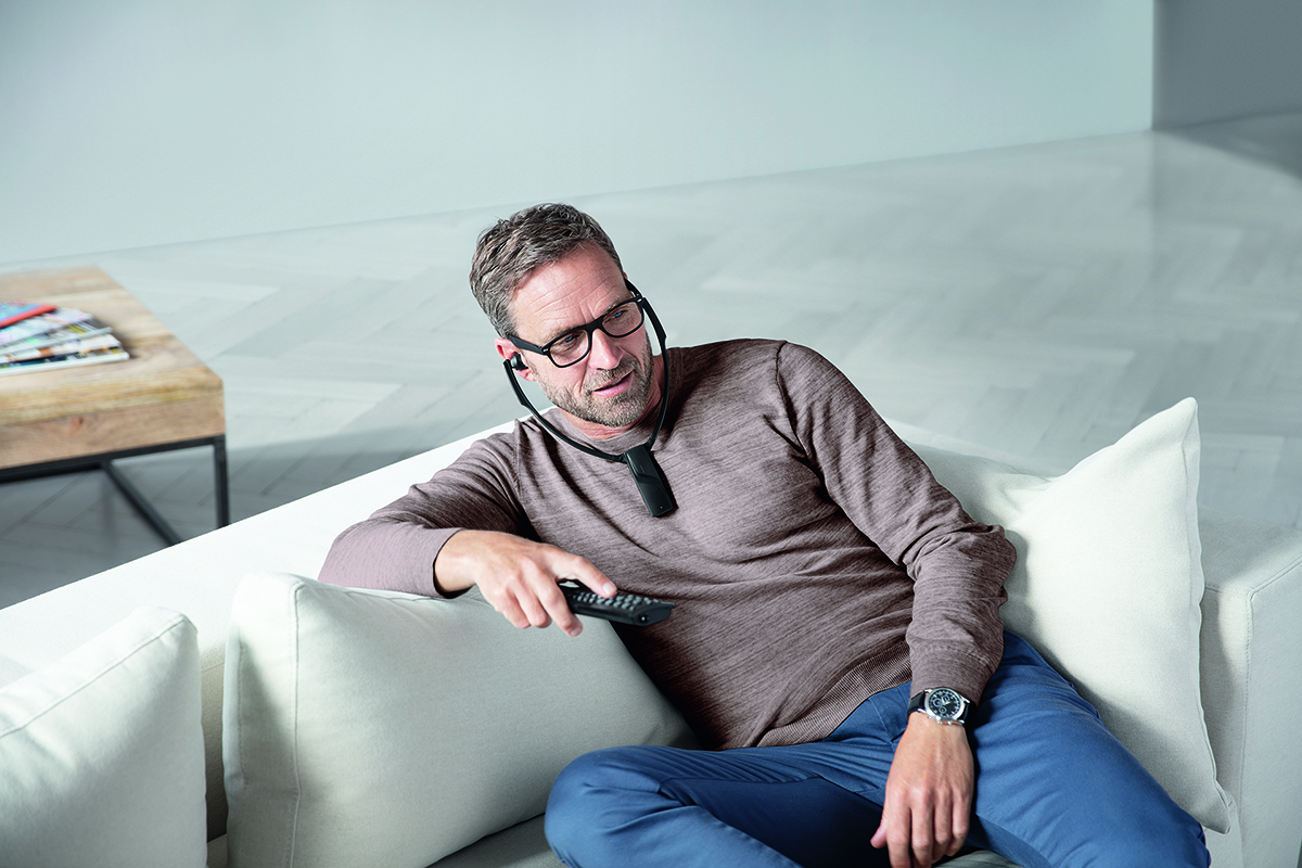 A man sitting on a white couch, wearing glasses and a neckband device, holding a remote.
