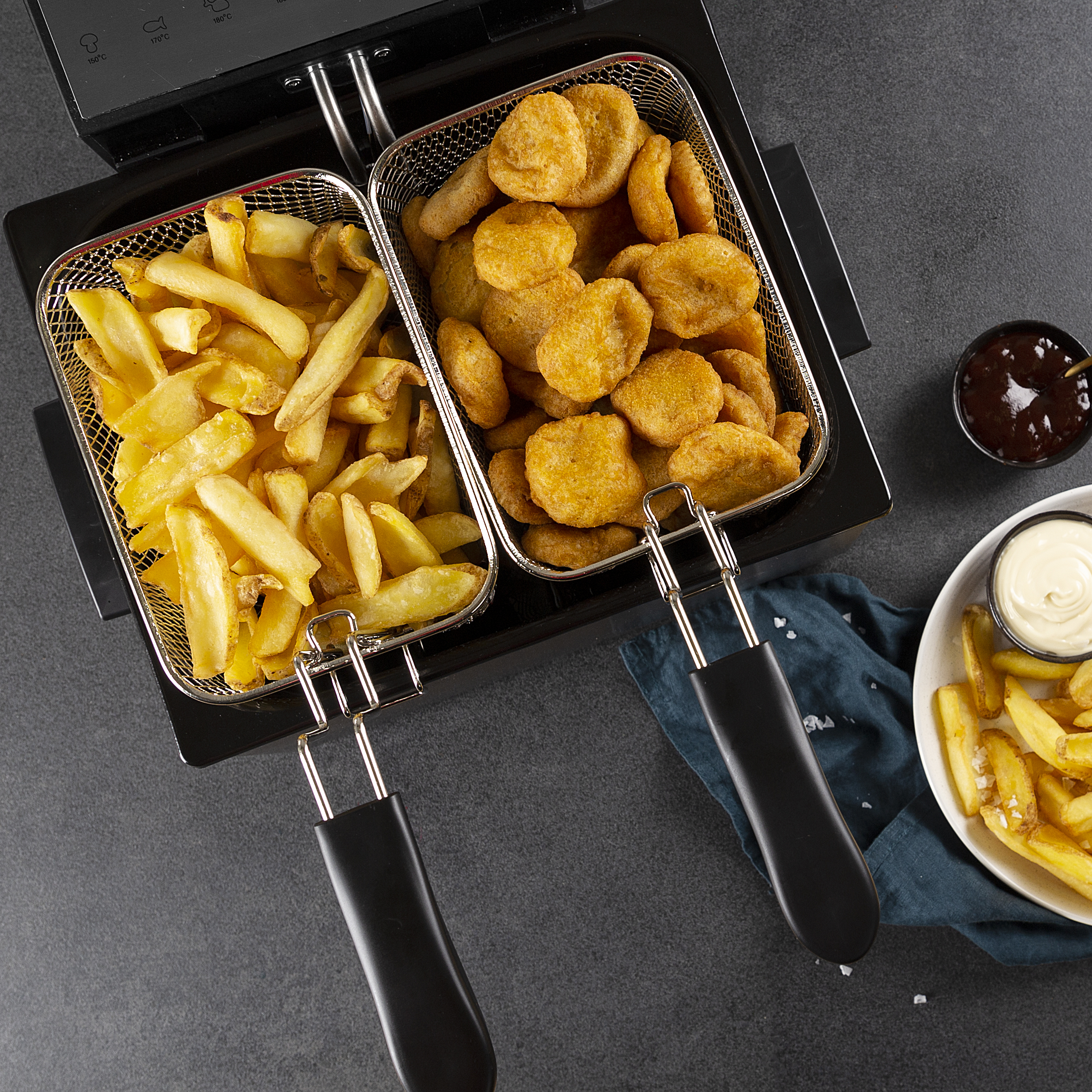 Zwei Körbe mit Pommes und Nuggets in einer Fritteuse, Beilagen daneben.