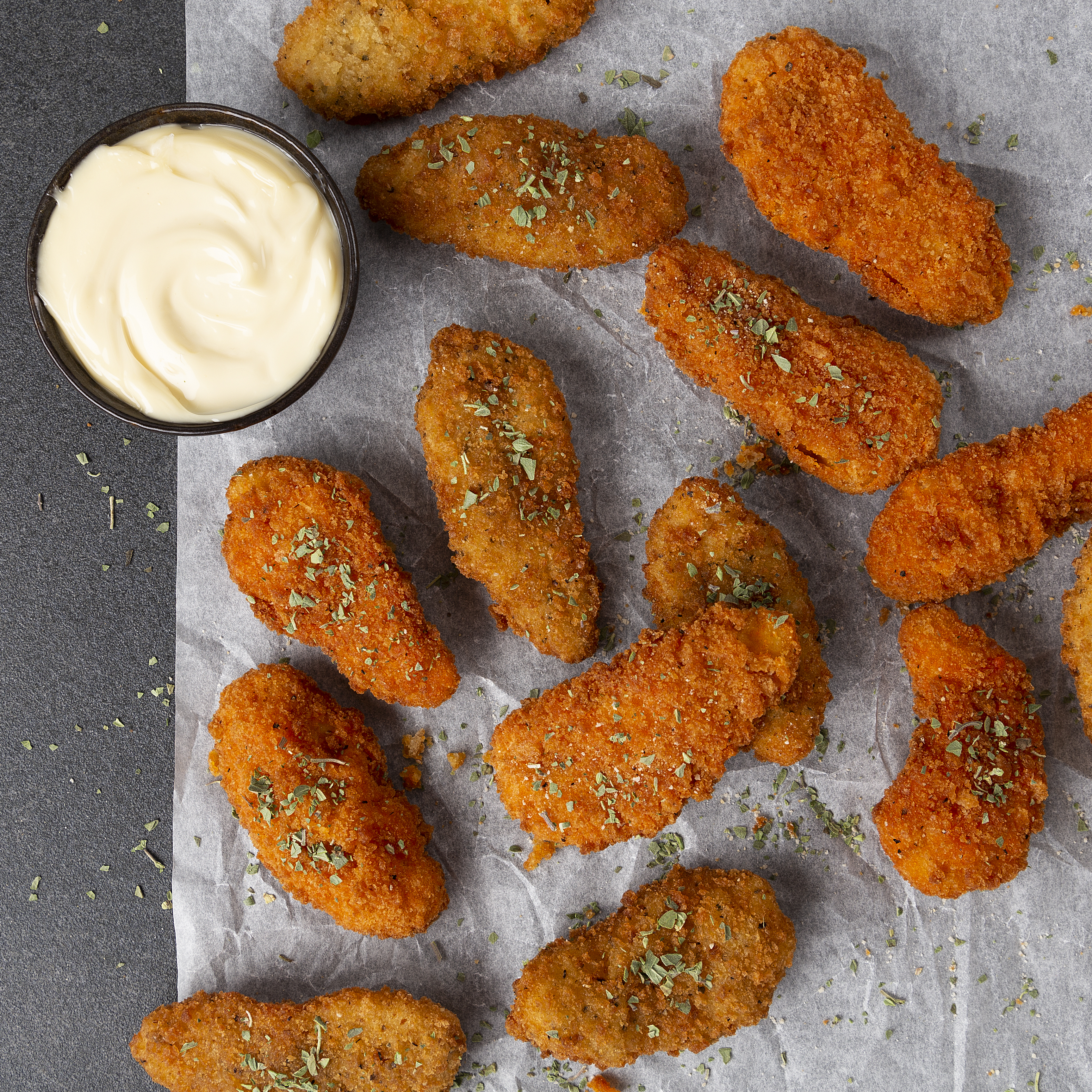 Frittierte Hähnchenstreifen mit Kräutern und eine Schüssel Mayo auf grauer Oberfläche.