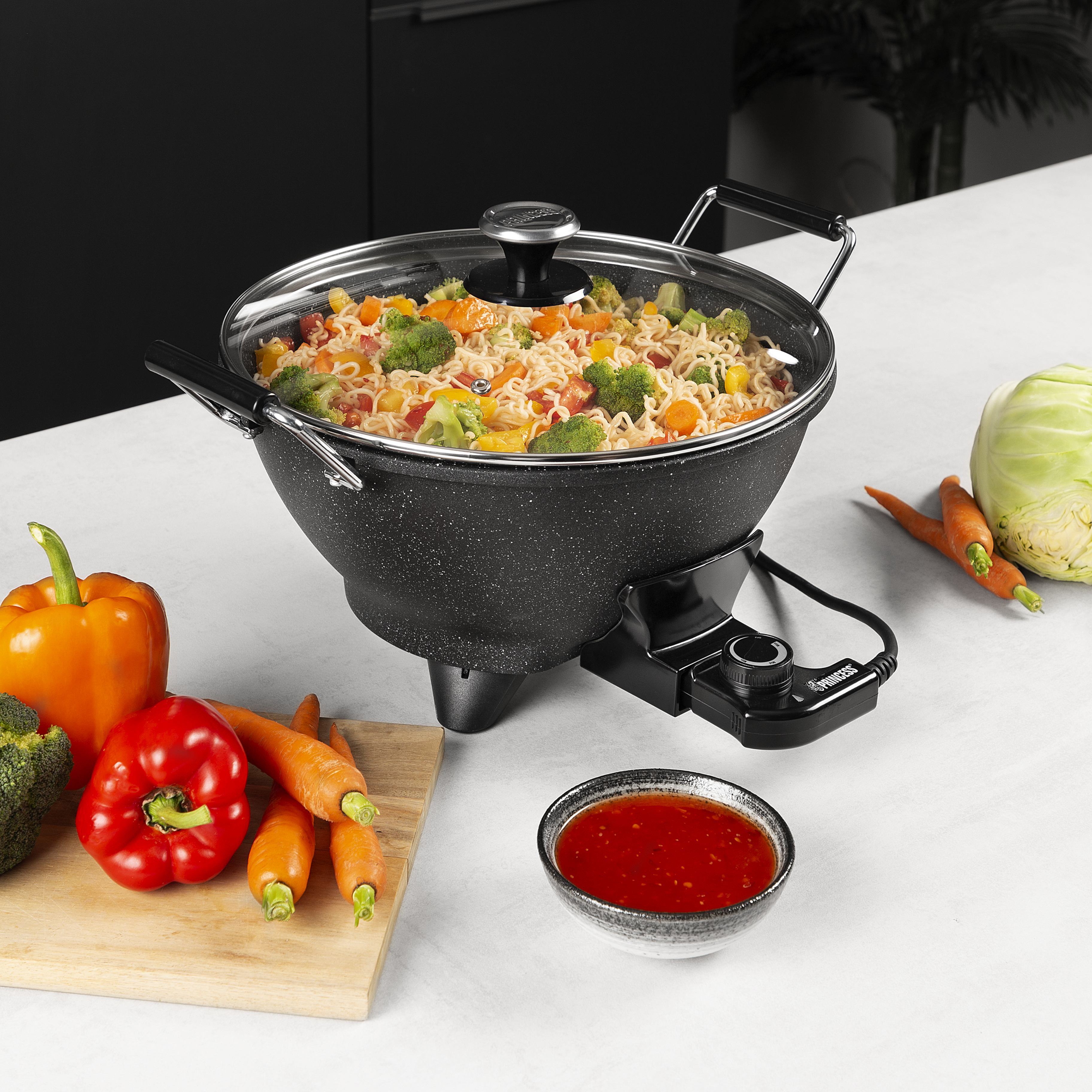A wok with cooked noodles and vegetables. Carrots, broccoli, and red pepper are on the table.