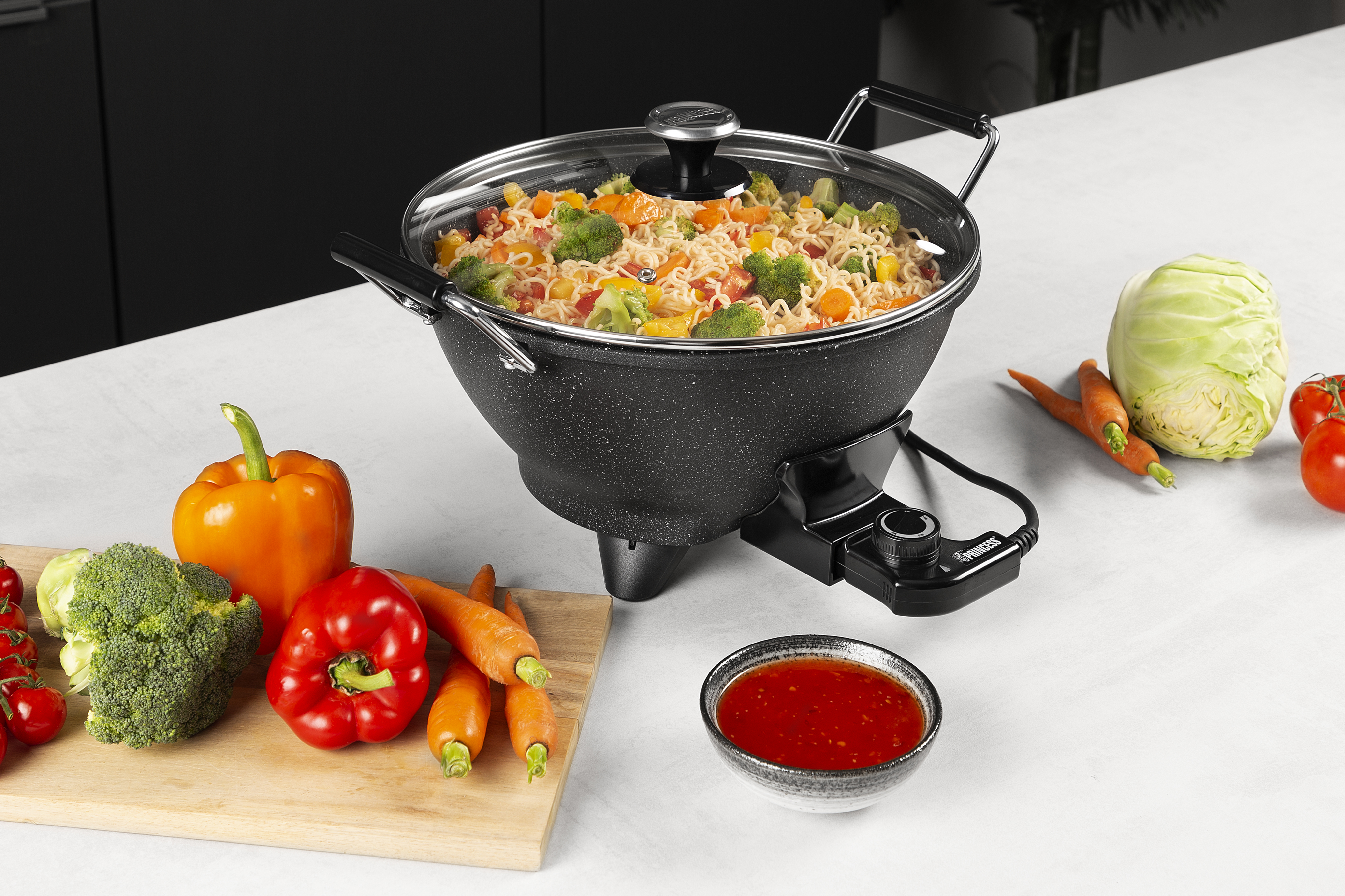 A wok with cooked noodles and vegetables, surrounded by fresh vegetables and sauce.