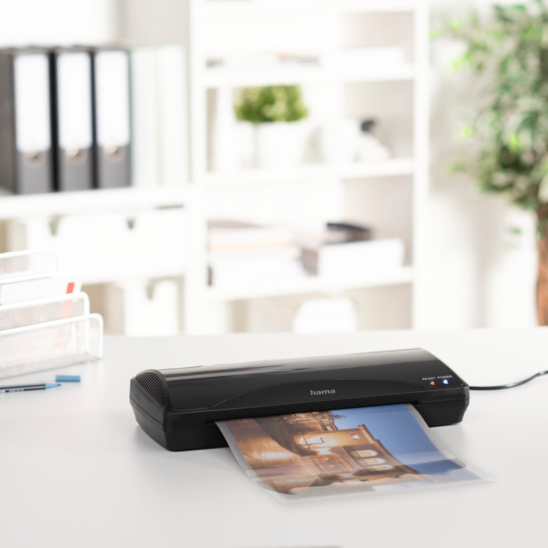 Black laminator with a photo emerging, on a white desk. Shelves and plants in the background.