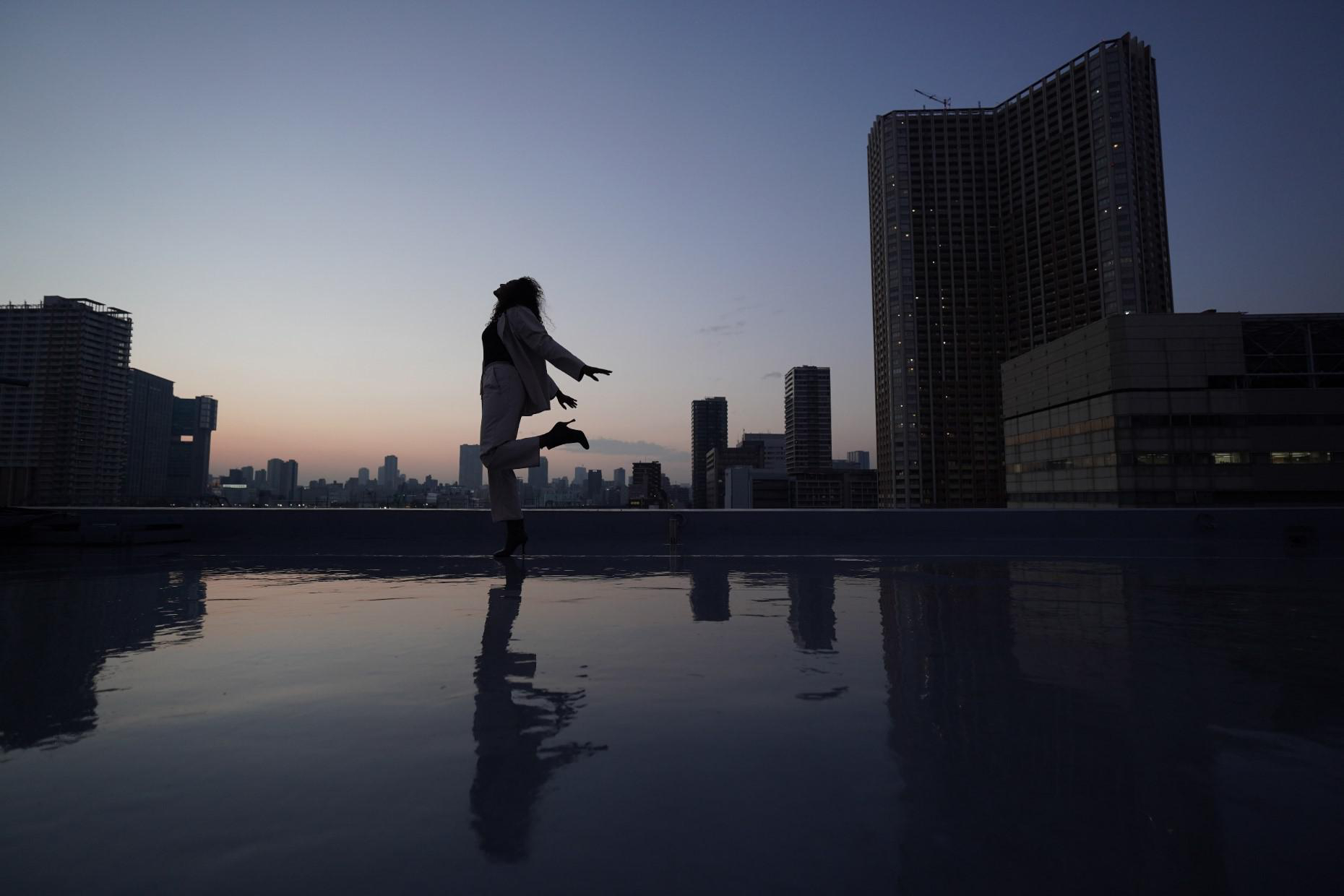Frau tanzt bei Dämmerung auf einem Dach, Silhouette vor Skyline. Dunkle, reflektierende Oberfläche.