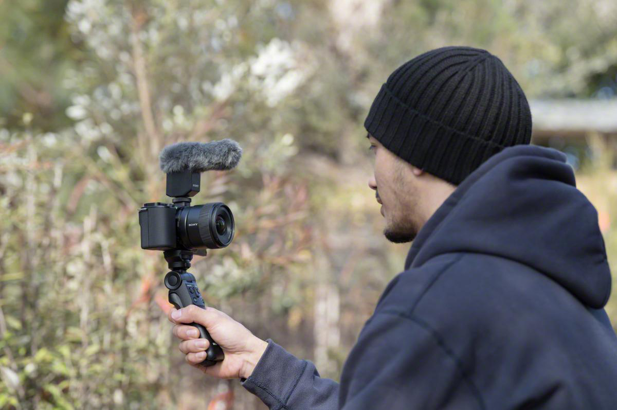 Ein Mann hält eine Kamera mit Mikrofon auf dem Dach und nimmt draußen ein Video auf, mit Bäumen im unscharfen Hintergrund.