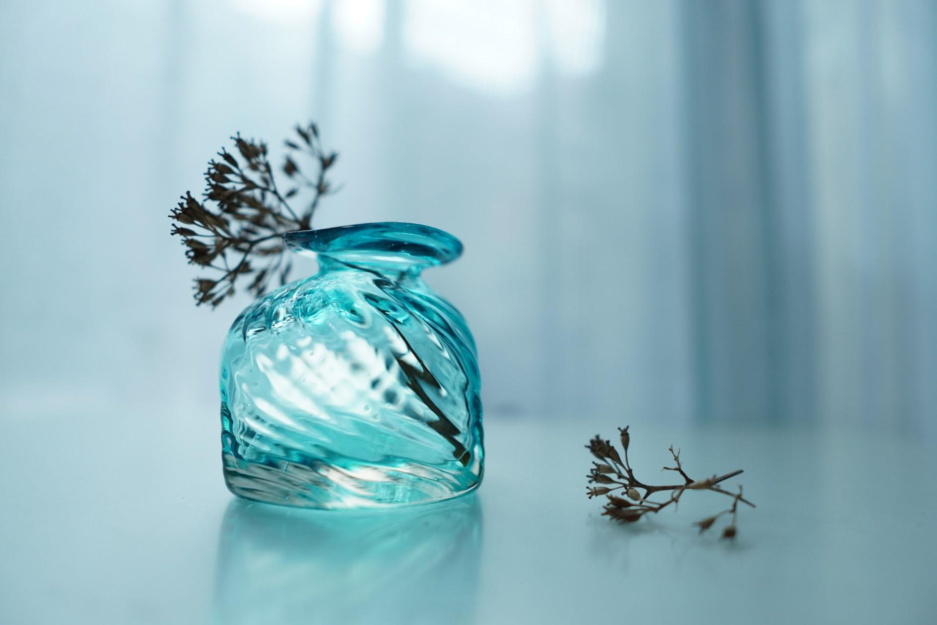 Eine türkisfarbene Glasvase mit getrockneten Blumen. Ein Zweig Blumen liegt auf dem Tisch.