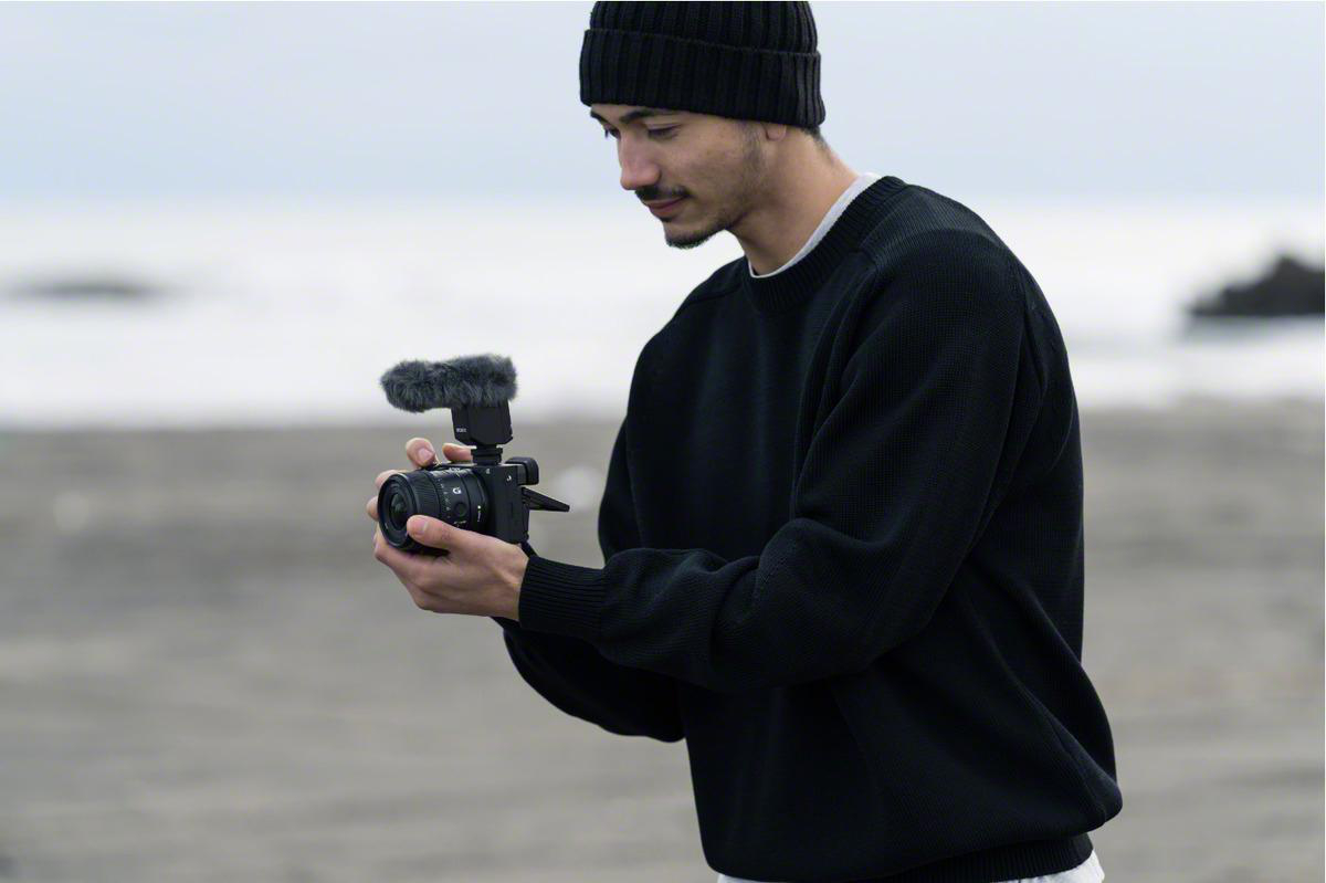 Ein Mann filmt mit einer Kamera mit Mikrofon. Der Hintergrund ist ein bewölkter Strand.