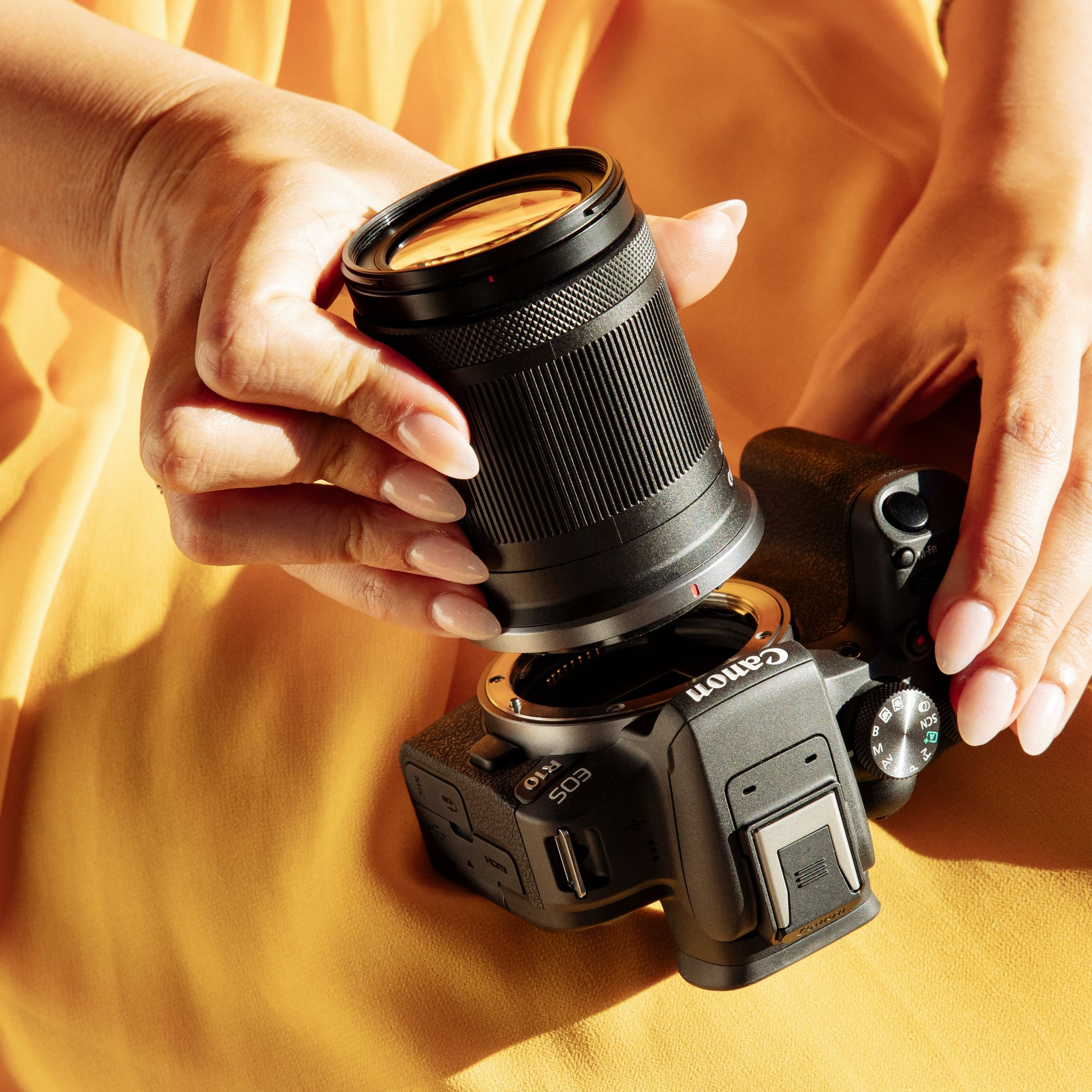 Mani che tengono un obiettivo sopra una fotocamera Canon EOS R10, su sfondo giallo.