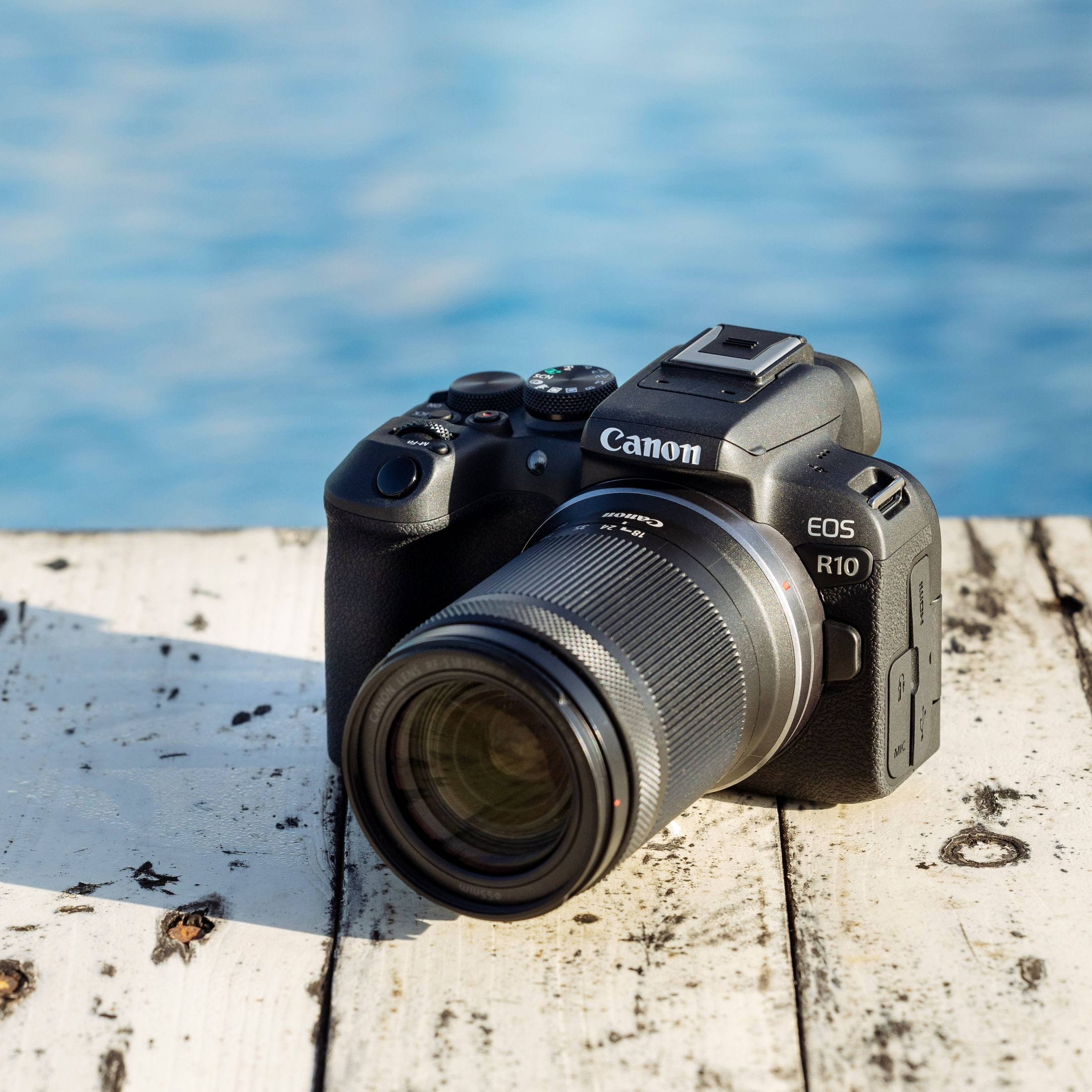 Una fotocamera Canon EOS R10 nera con obiettivo è su una superficie di legno bianca, acqua blu sullo sfondo.