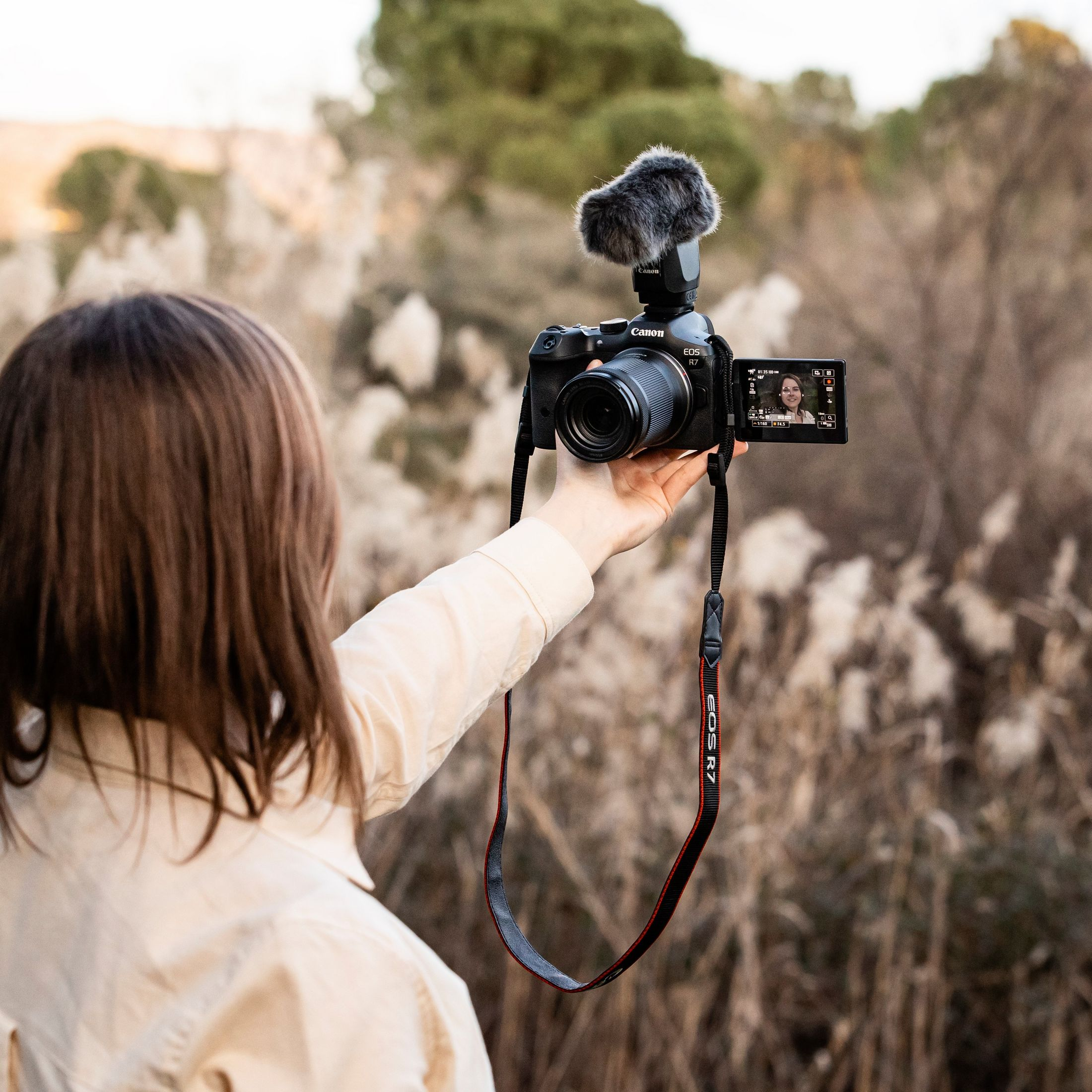Una persona tiene una fotocamera con un microfono. La fotocamera mostra un video della persona. Lo sfondo è erba alta.