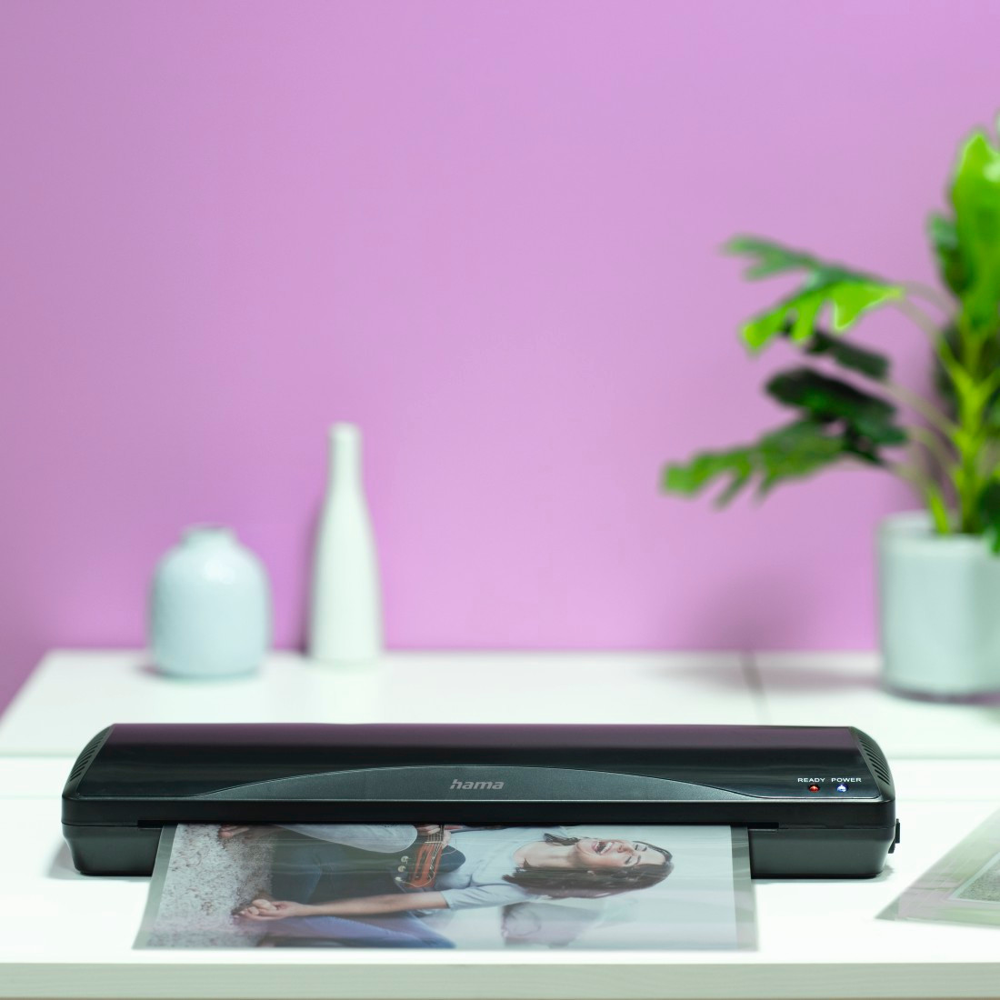 A black laminator with a photo in it, a pink wall, and a green plant are on a white table.