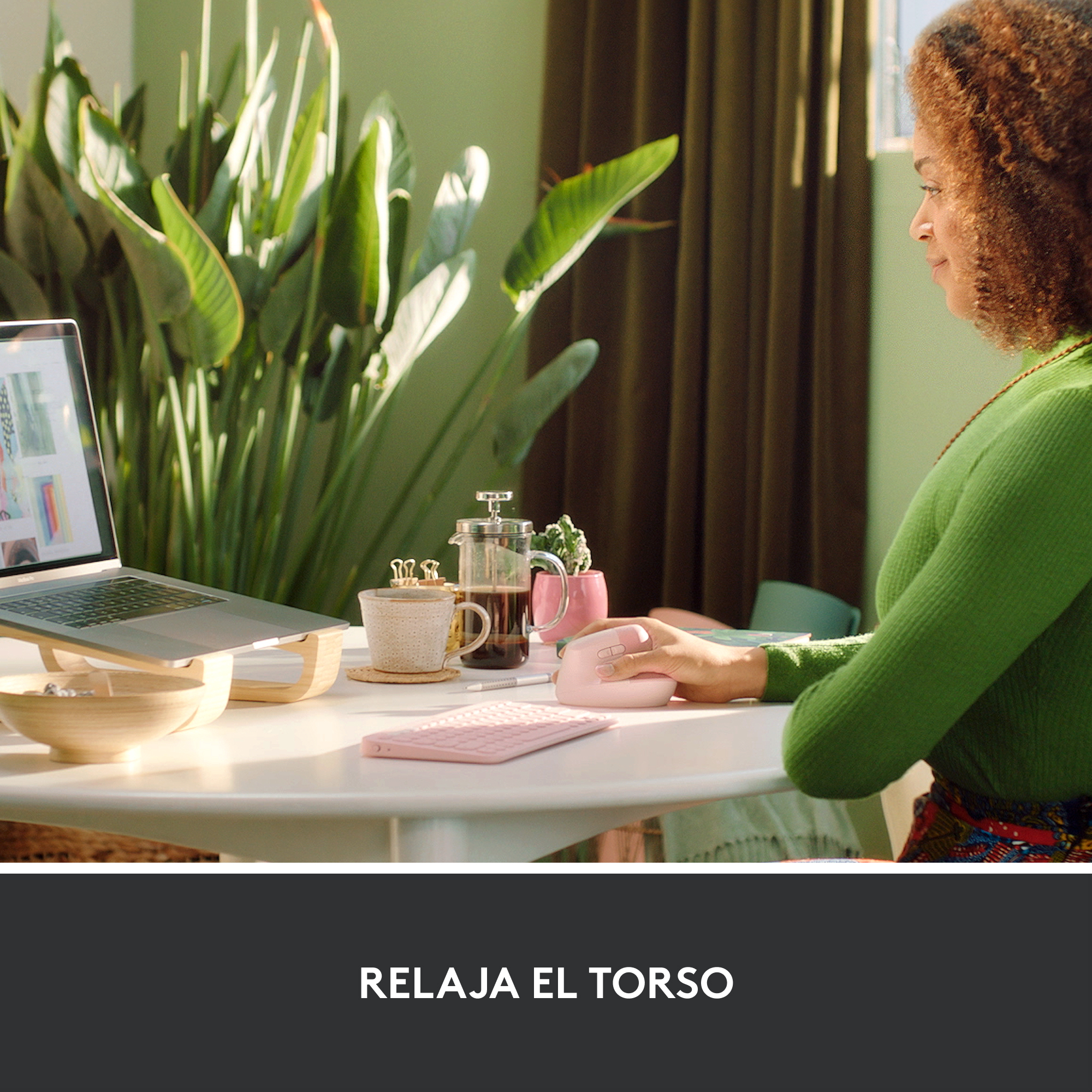 Mujer con suéter verde, trabajando en portátil. Teclado, ratón y taza de café rosas en mesa.